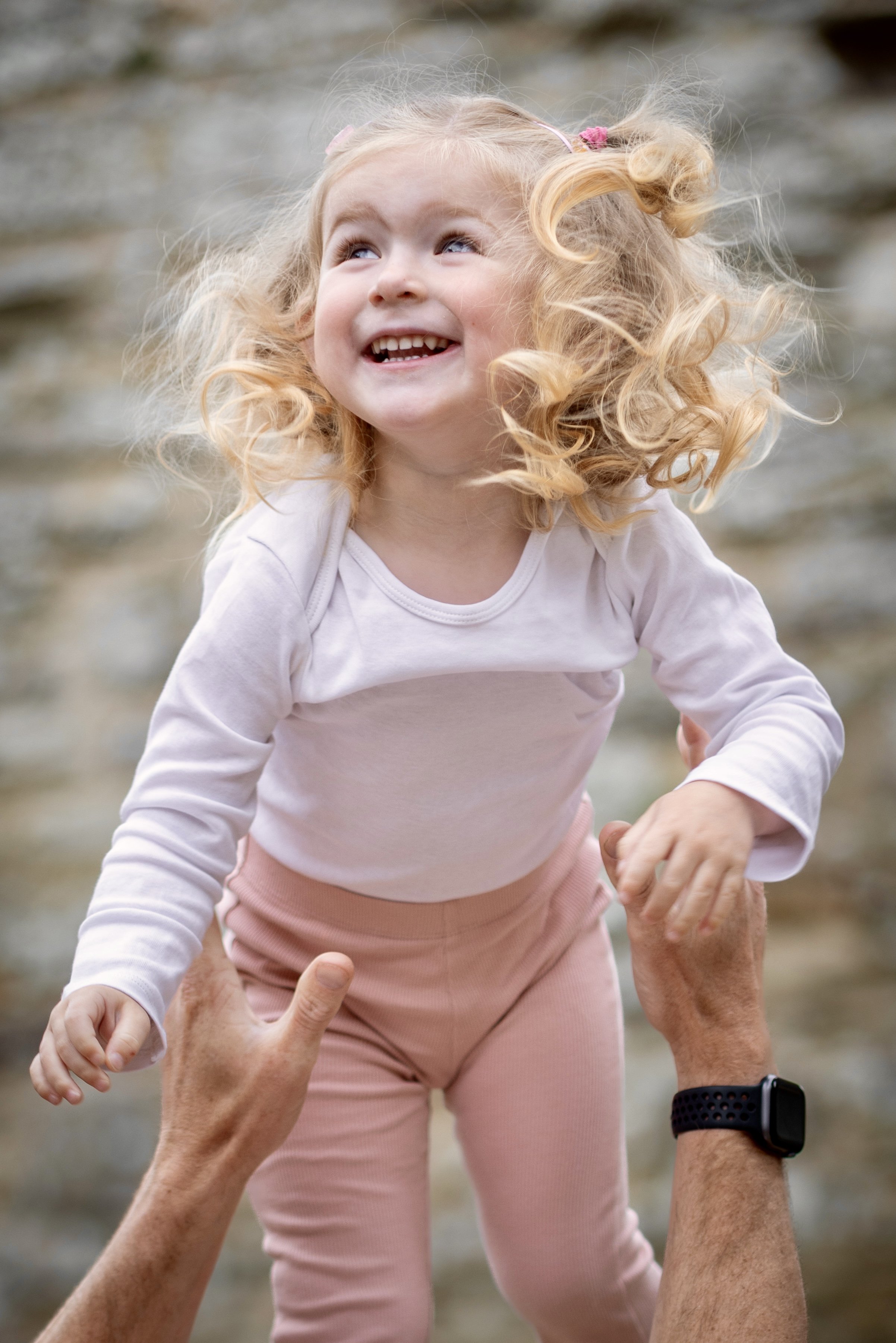 A young girl with blonde, curly hair wearing a pink long-sleeve shirt smiling and looking up while being held up by an adult outdoors near a beach or rocky area.