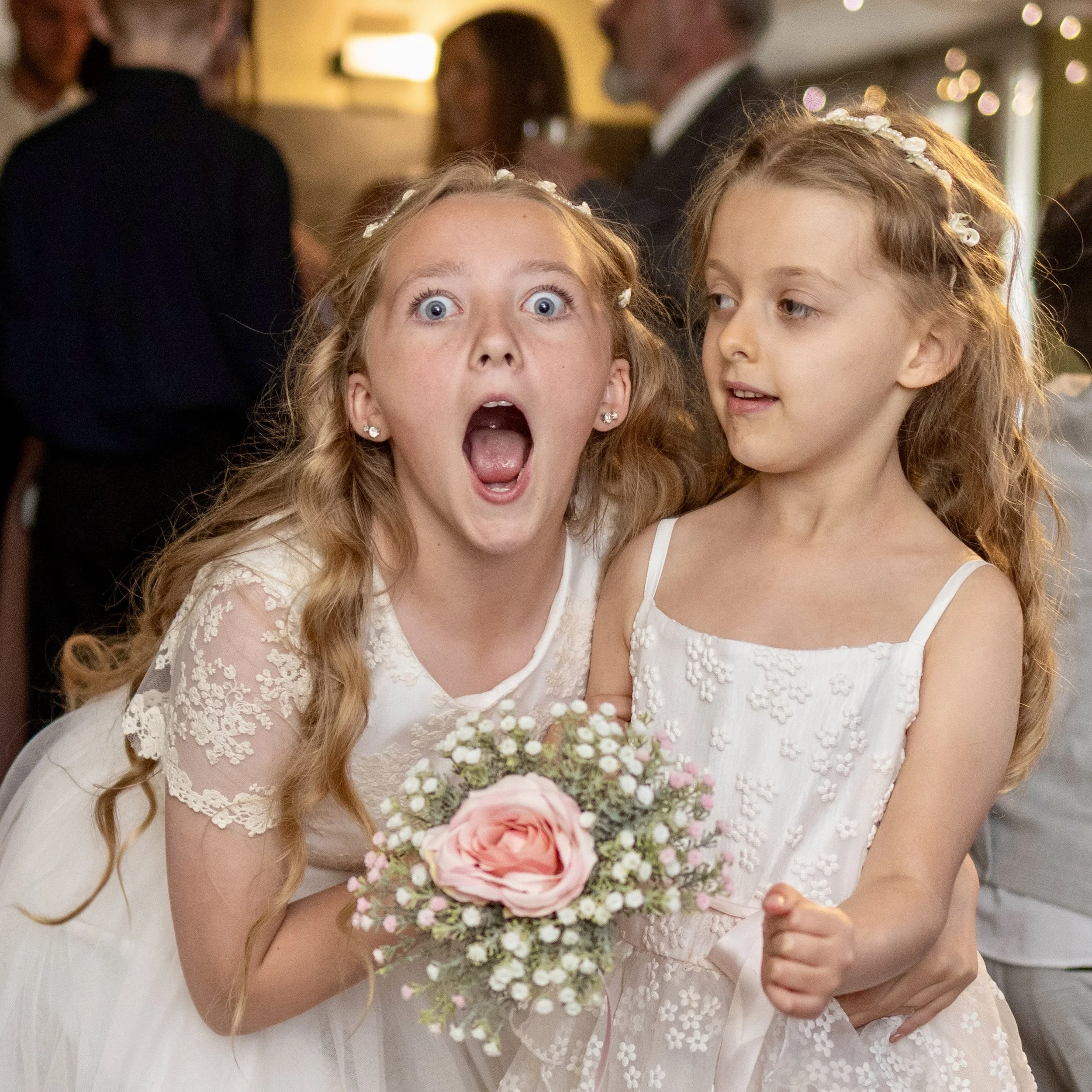 Two young girls in white dresses at a celebration, one with mouth wide open and holding a bouquet of pink and white flowers, while the other looks on with a smile.