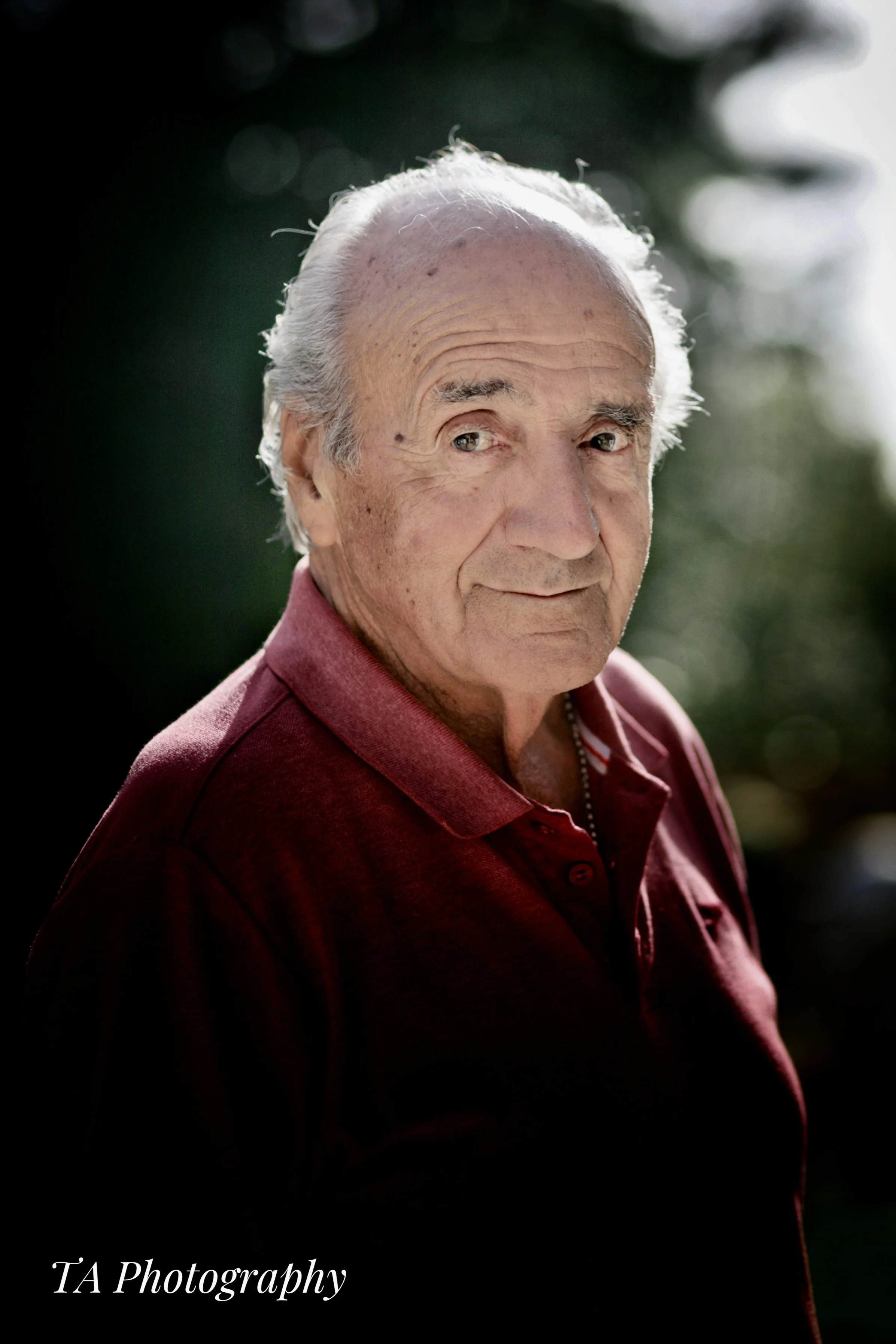 A close-up of an elderly man with white hair, wearing a dark red collared shirt, standing outdoors with a blurred background of green trees and sunlight.
