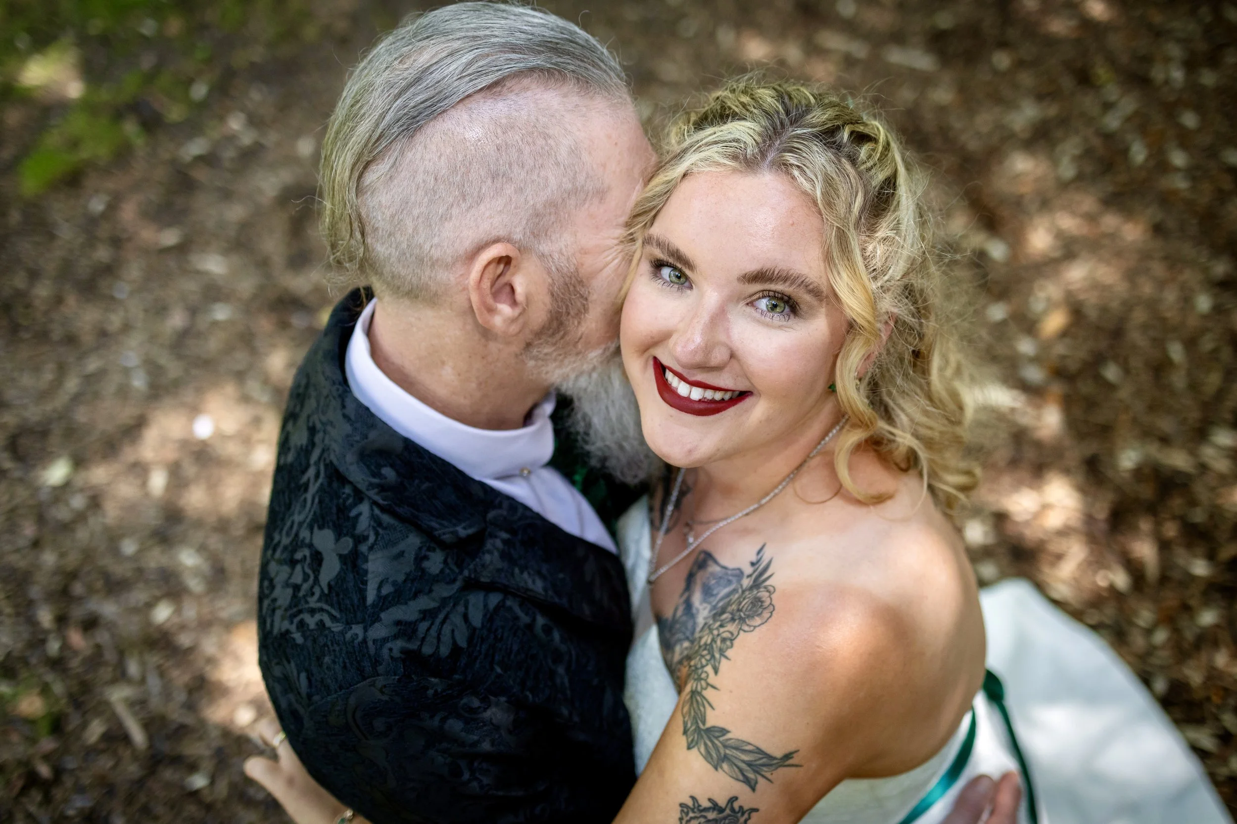 A smiling woman with curly blonde hair and tattoos on her shoulder, wearing a white dress with green accents, is being embraced from behind by a bearded man with short blonde hair and a shaved side, dressed in a dark patterned suit. They are outdoors
