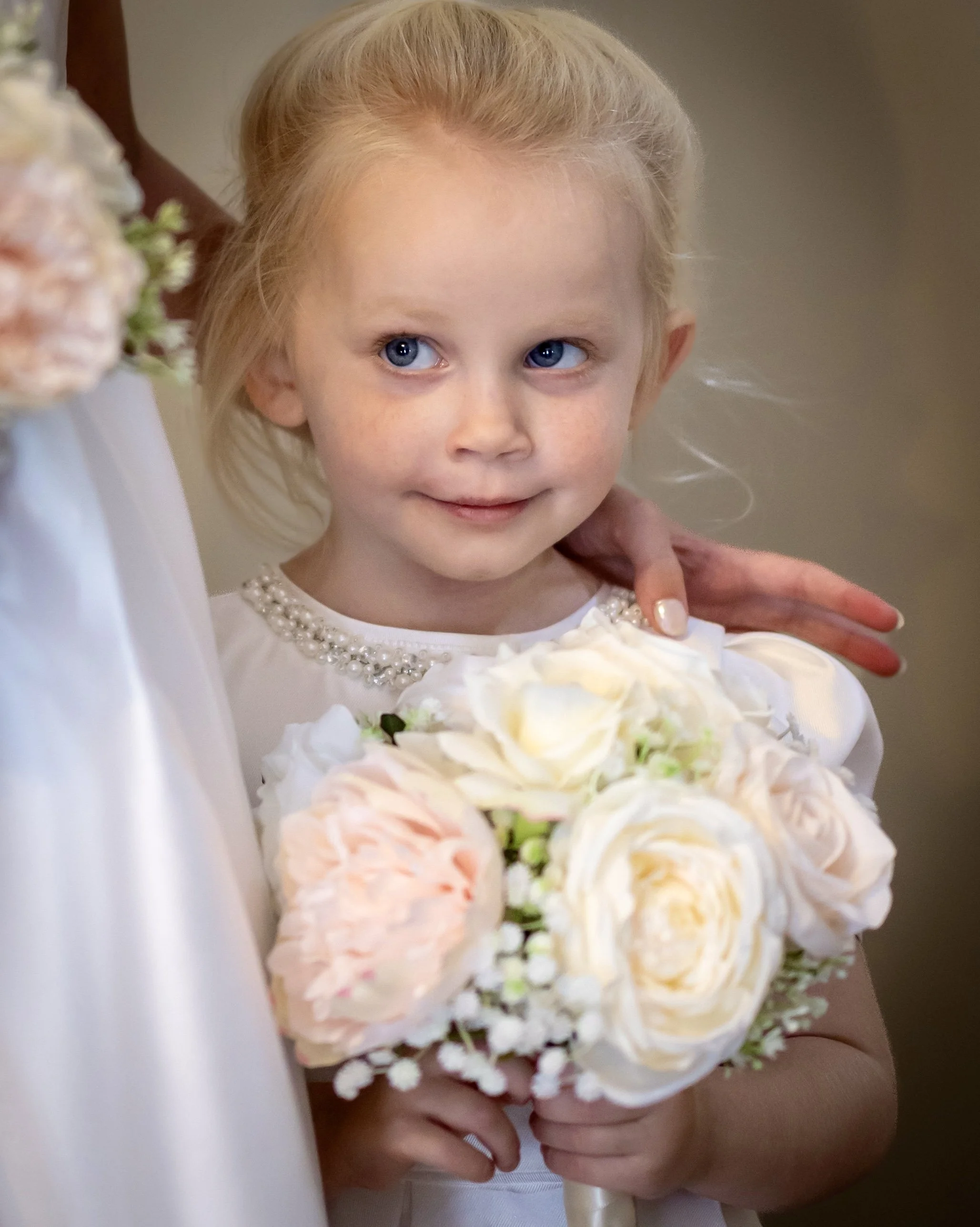 Close-up of a young girl with blonde hair and blue eyes holding a bouquet of white and peach roses, with a pearl necklace, during what appears to be a special occasion.