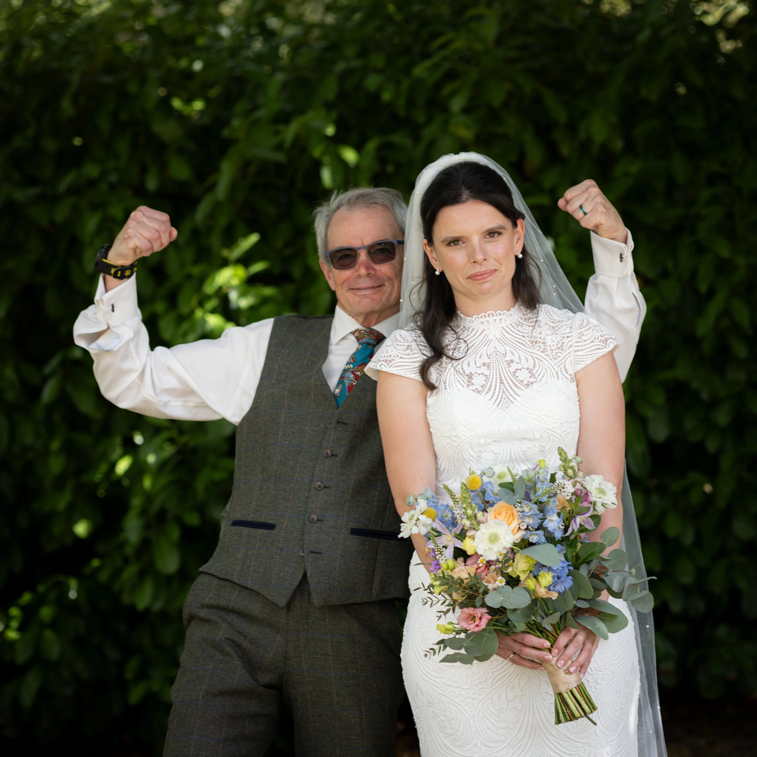 A bride holding a bouquet poses with a man in a suit vest, who is making a strong arm pose.