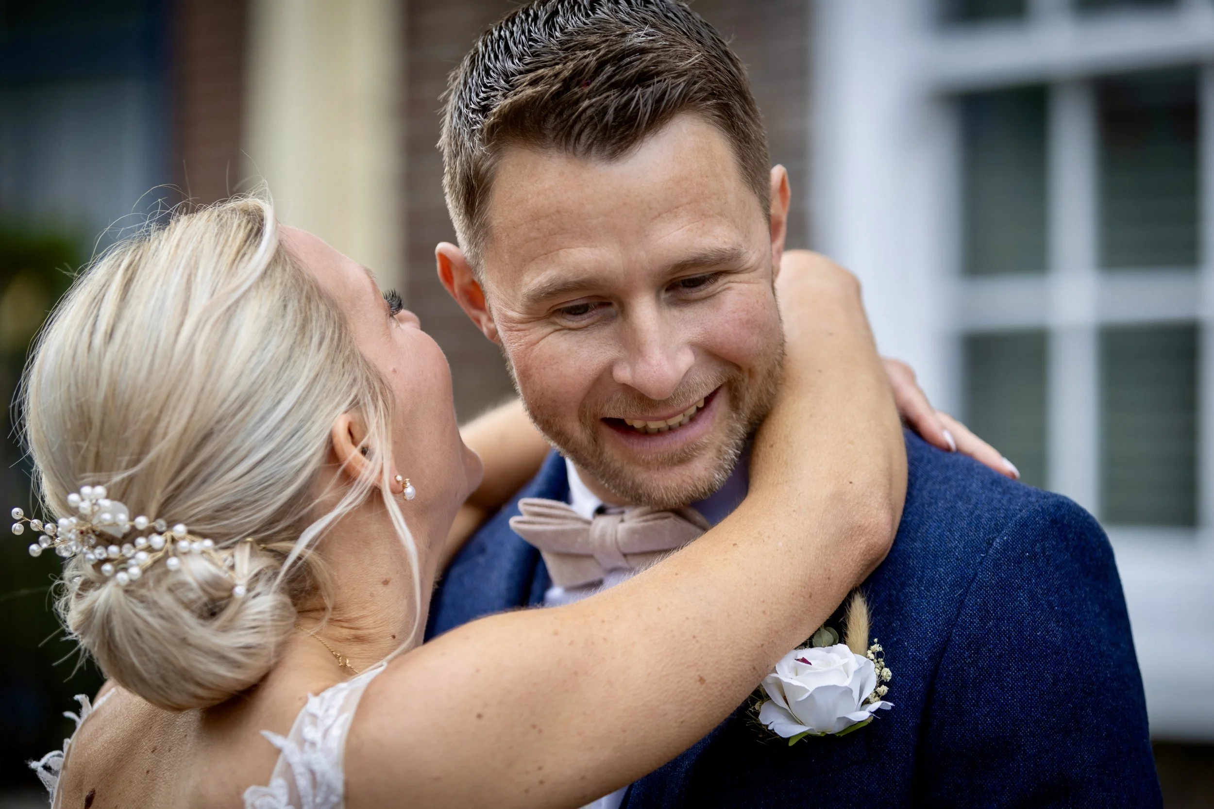 A man and a woman embrace, smiling affectionately. The woman has blonde hair styled with pearls, and the man has short brown hair with a beard. They are outside, with blurred houses in the background, suggesting a celebratory or romantic moment.