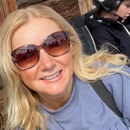 A woman with blonde hair wearing sunglasses and a blue shirt, smiling at the camera, sitting outdoors with a person in a black jacket and helmet in the background.