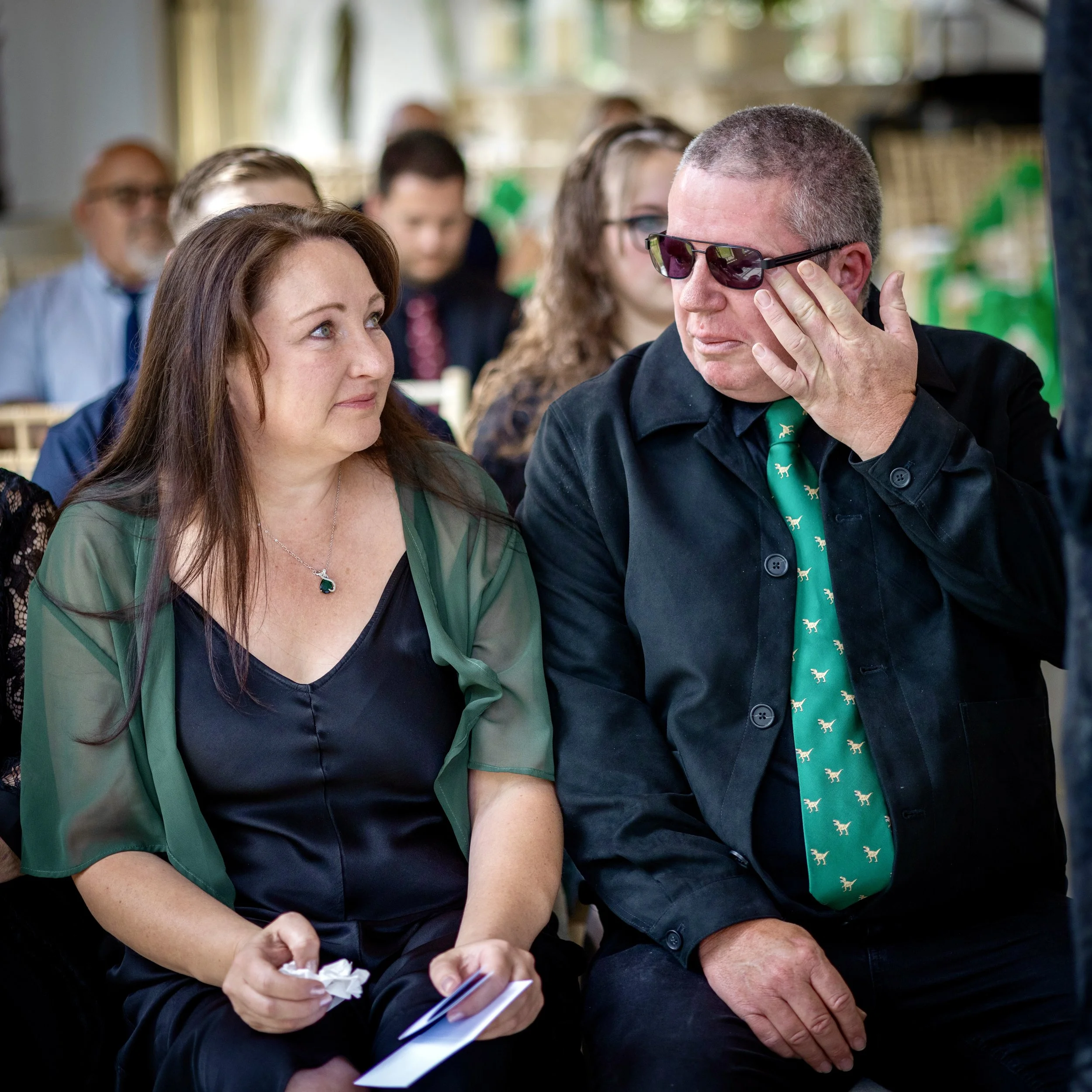 A man wearing sunglasses and a woman with long red hair are engaged in a conversation at an indoor gathering with other people in the background. The woman is holding a piece of paper and the man is touching his face while listening.