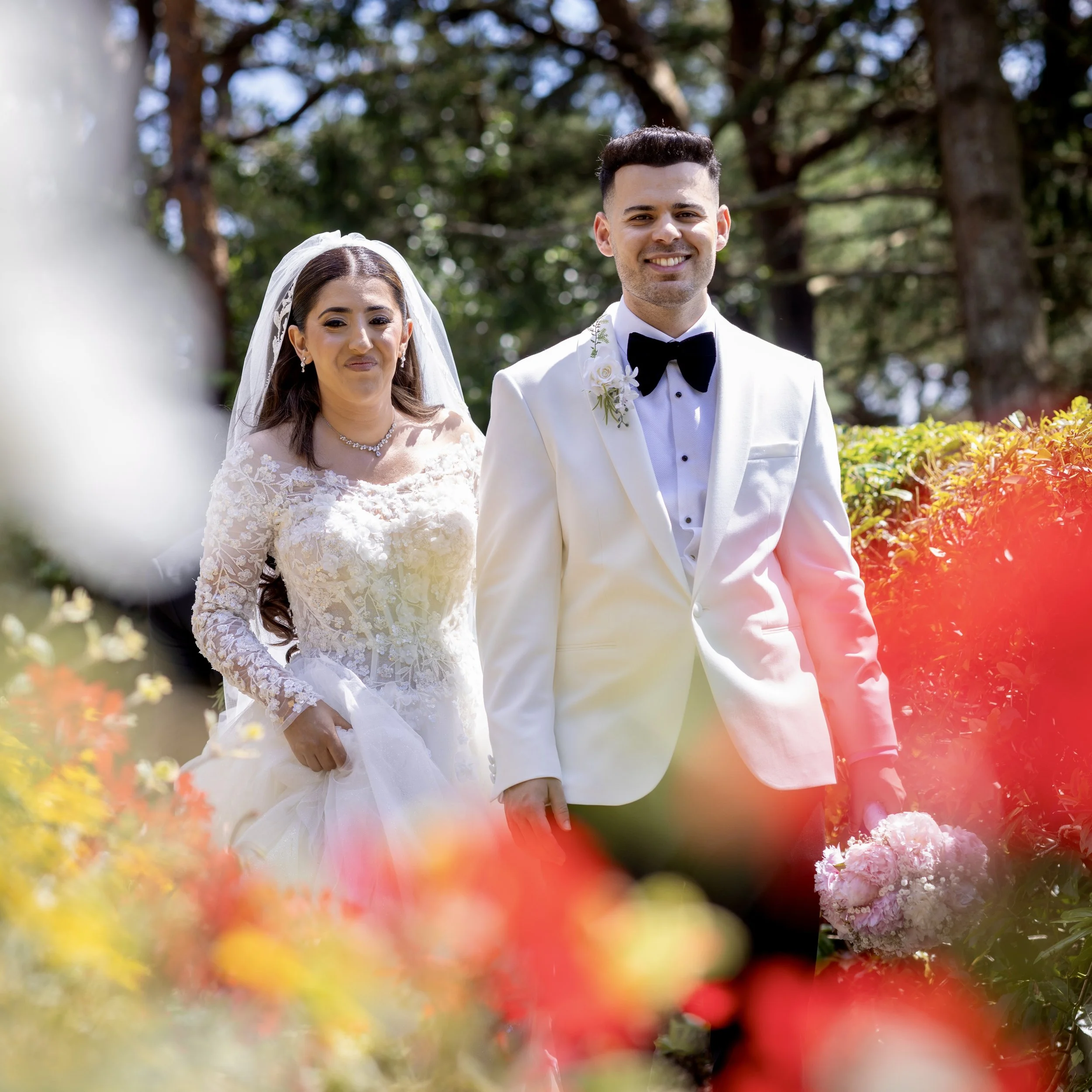 A bride and groom walking outdoors on their wedding day, surrounded by colorful flowers and greenery.