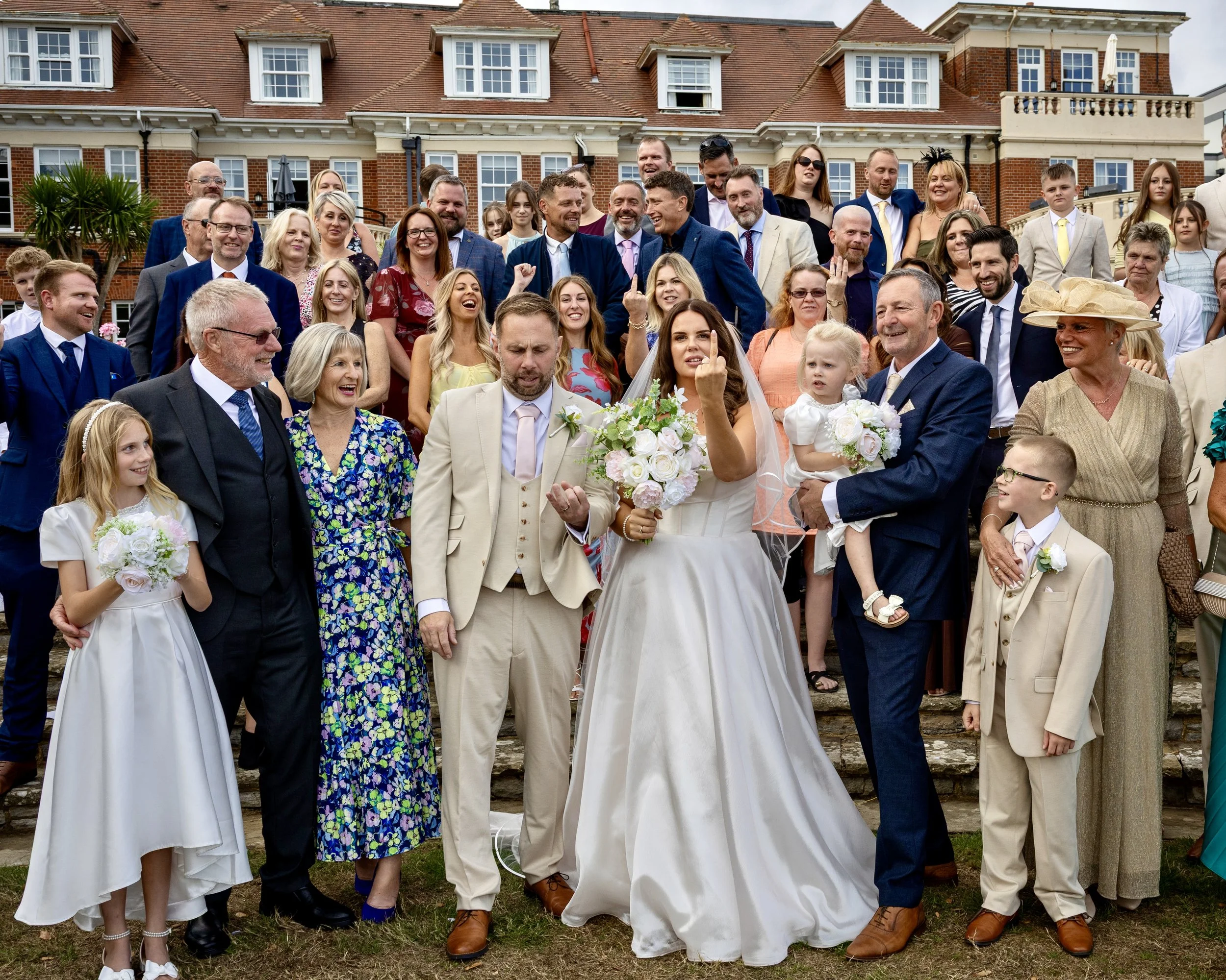 A large group of people, including men, women, and children, gathered outside in front of a brick and white building, celebrating a wedding. The bride, dressed in a white gown, is holding a bouquet of flowers and making a rude hand gesture. The group