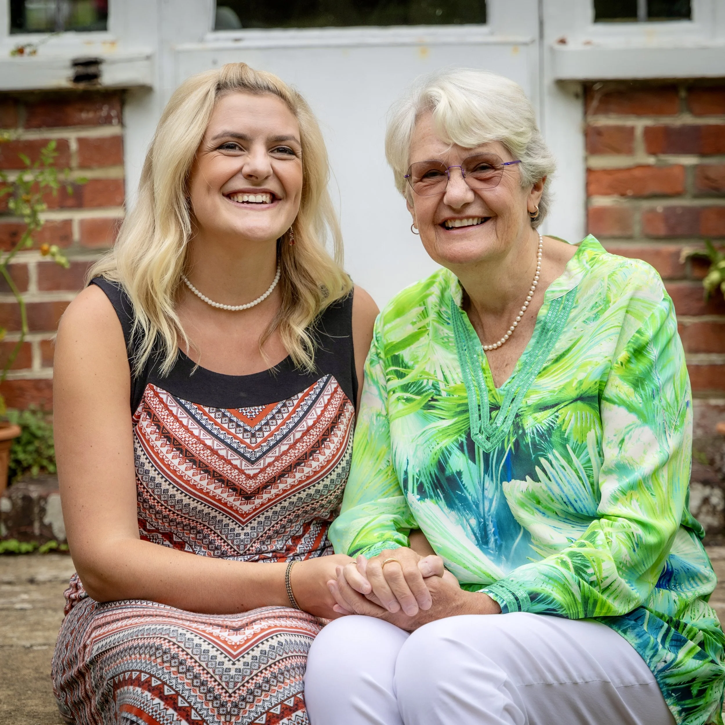 Two women sitting outdoors against a brick wall, smiling, with their hands clasped together.