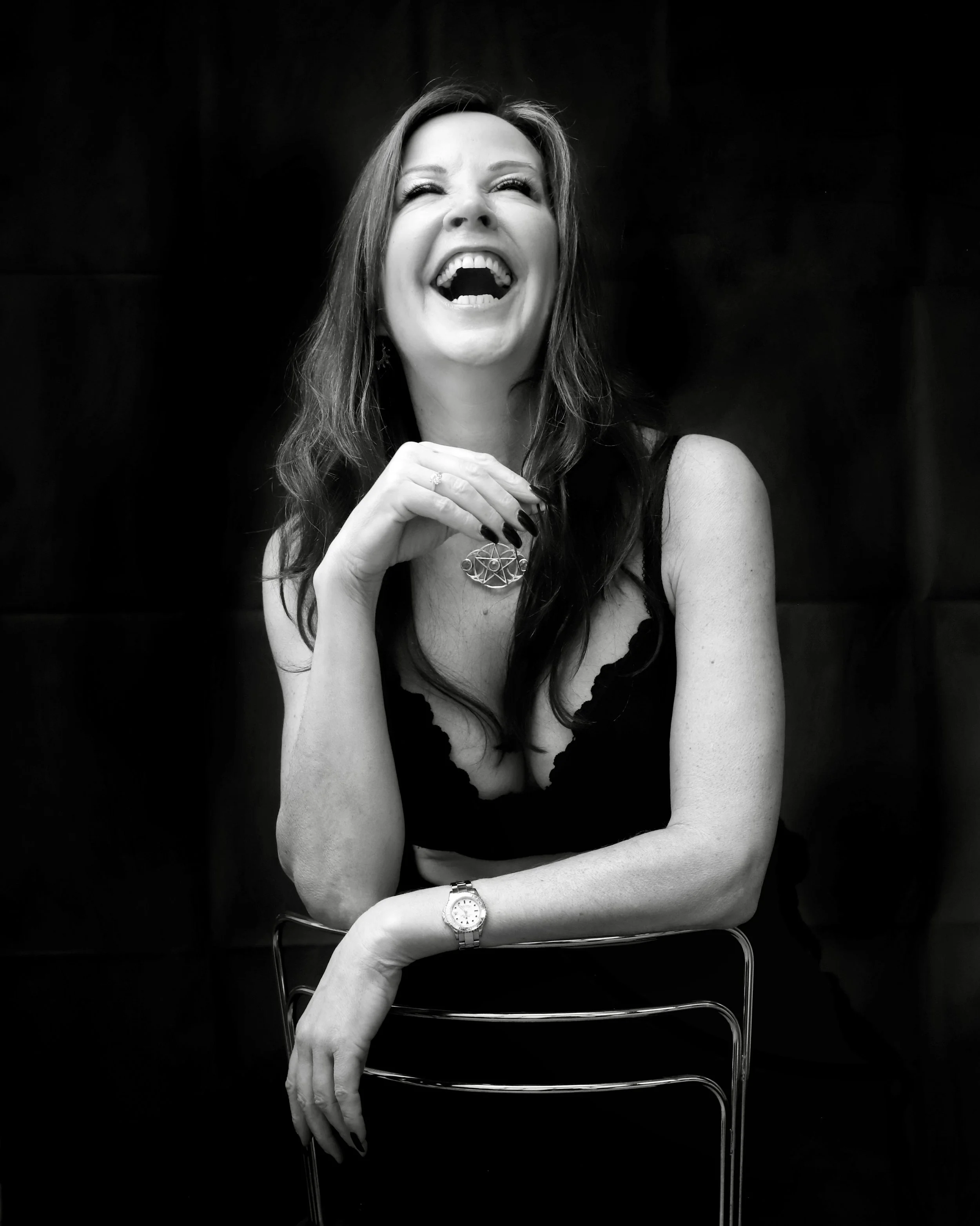 A woman with long wavy hair laughing with her eyes closed, wearing a watch on her left wrist, a ring, and a necklace with a pendant. She is sitting in front of a dark background, with her left arm resting on a chair. The image is in black and white.