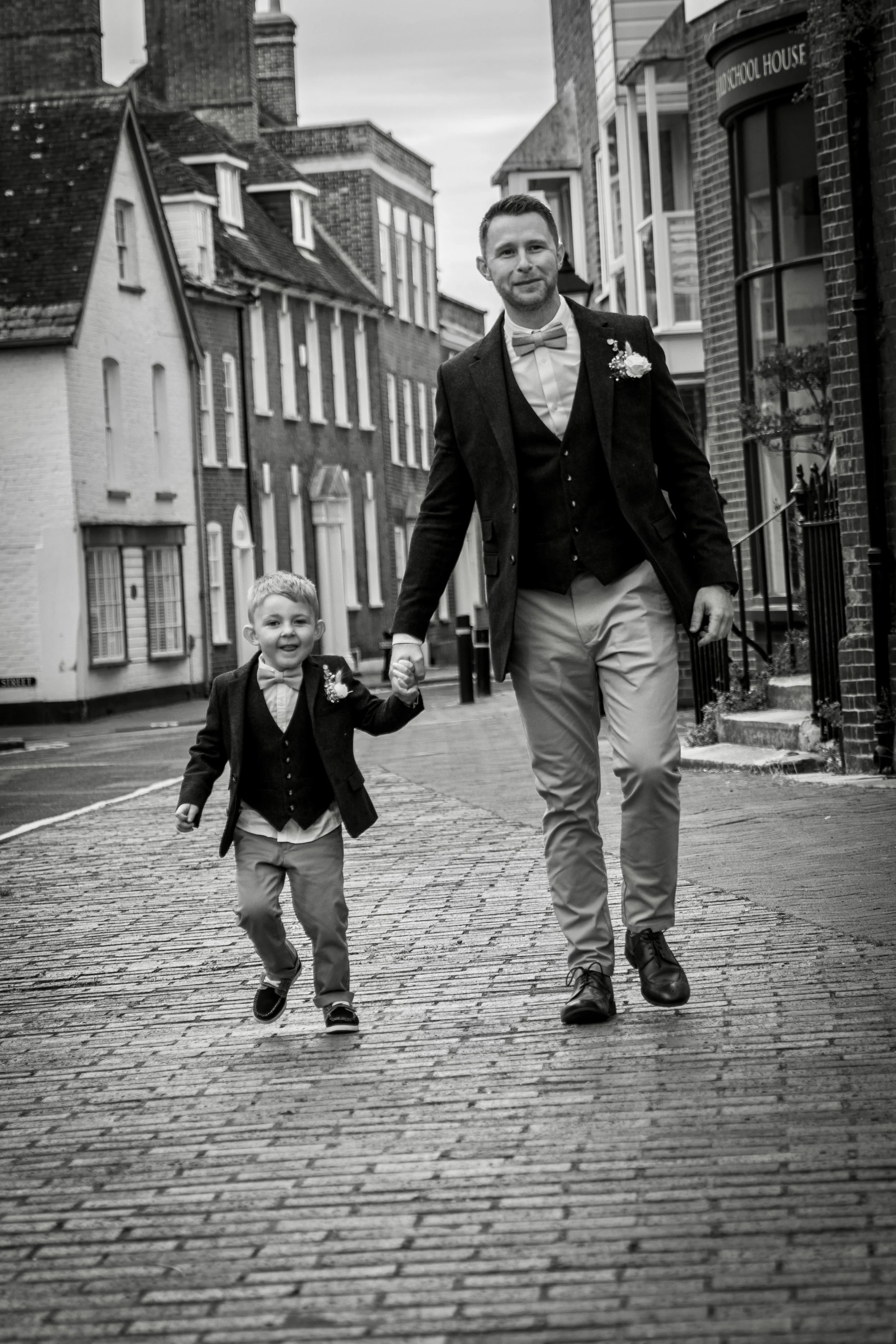 Black and white photo of a man in a suit and bow tie and a young boy in a suit and bow tie walking hand in hand on a cobblestone street with historic buildings in the background.