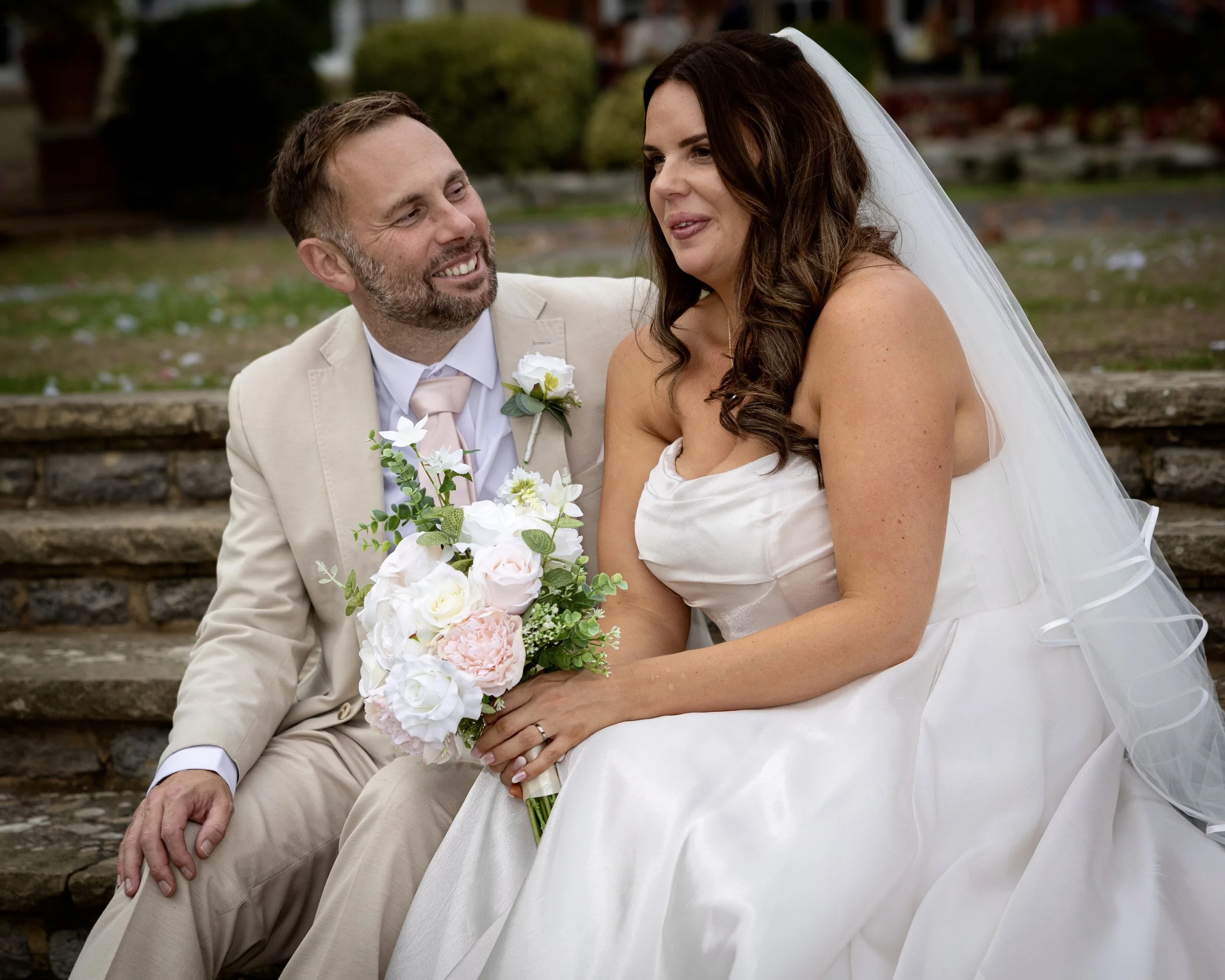 A smiling groom and bride sitting on outdoor stone steps during their wedding, with the groom in a beige suit and the bride in a white strapless wedding dress holding a bouquet of white and pink flowers.