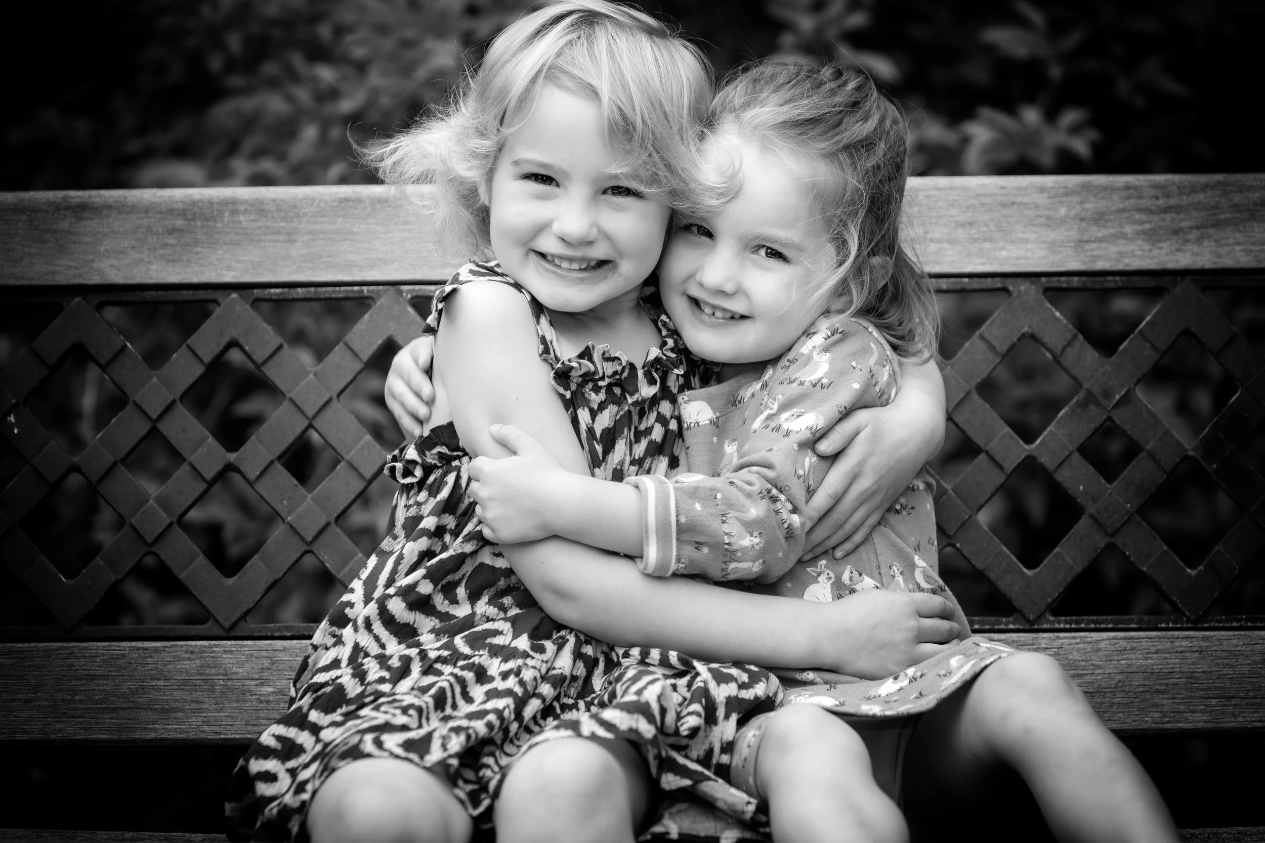 Two young girls with curly hair hugging and smiling while sitting on a park bench, in black and white.