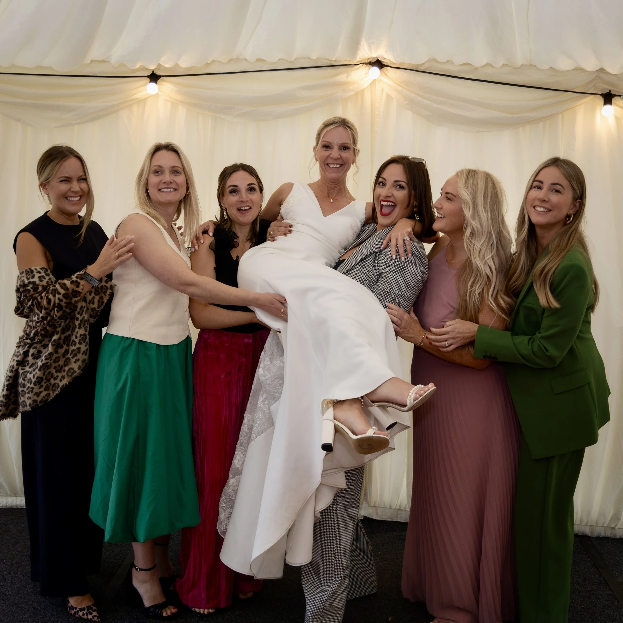 A group of seven women, smiling and posing together, with one woman being lifted by others at a happy event under a cream-colored tent with string lights.