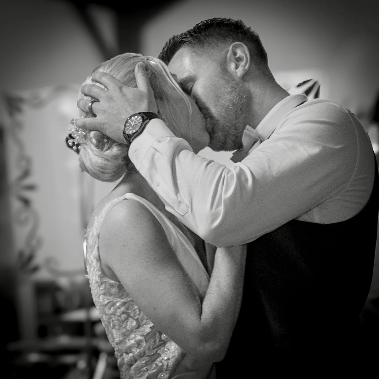 A black and white photo of a couple sharing a kiss, with the man holding the woman's head tenderly during a wedding celebration.