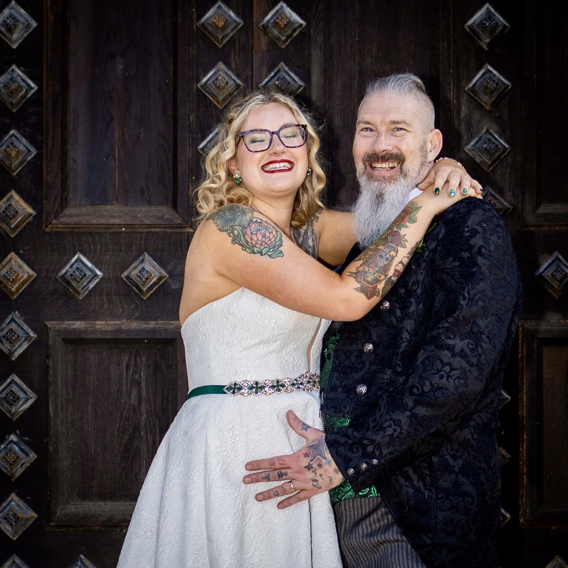 A smiling woman with curly blonde hair, glasses, and tattoos on her arms, wearing a white dress, hugging a man with a beard, tattoos, and grey hair, dressed in a black jacket with green accents, standing in front of a dark wooden background.
