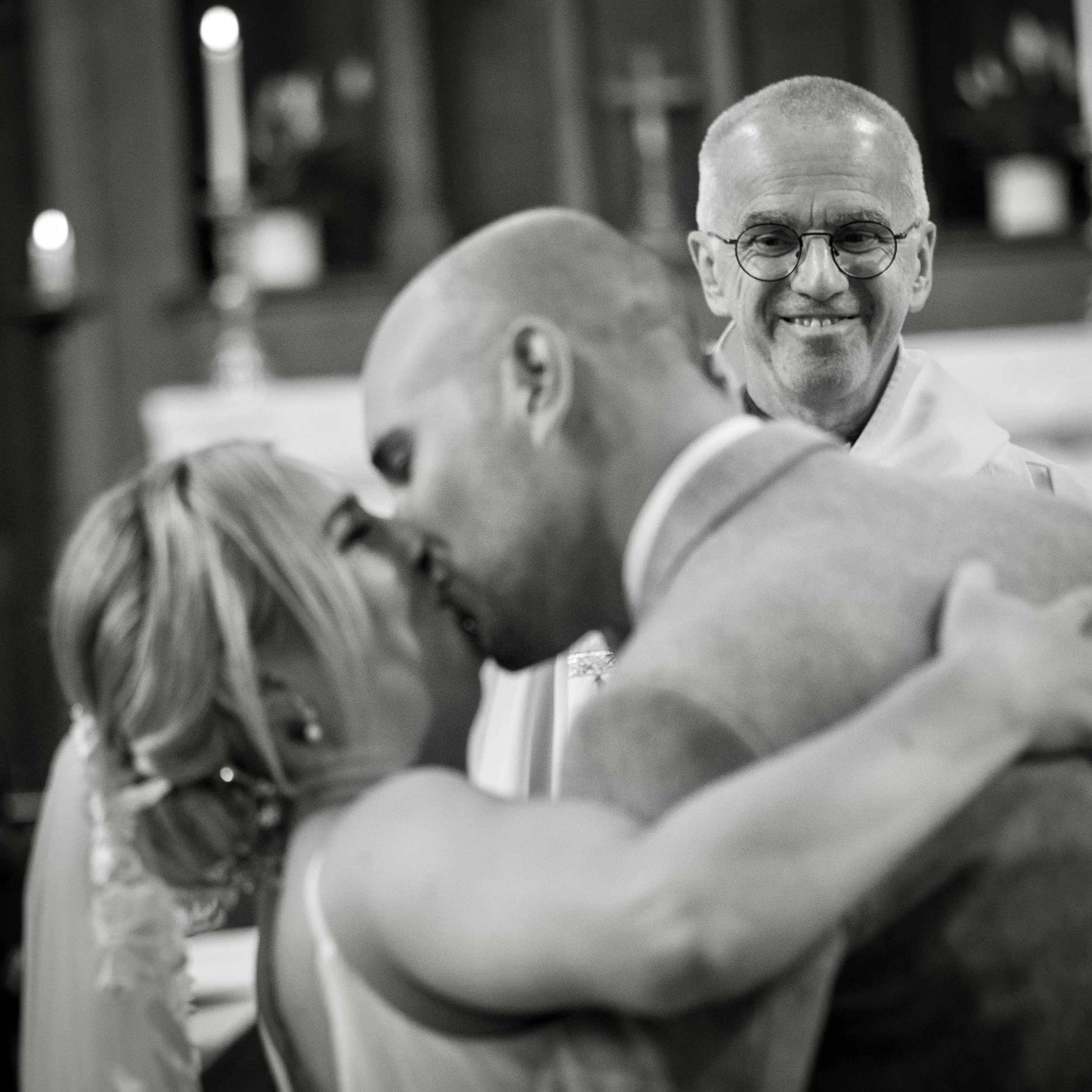 A wedding ceremony with a bride and groom sharing a kiss, while a smiling officiant looks on in the background.