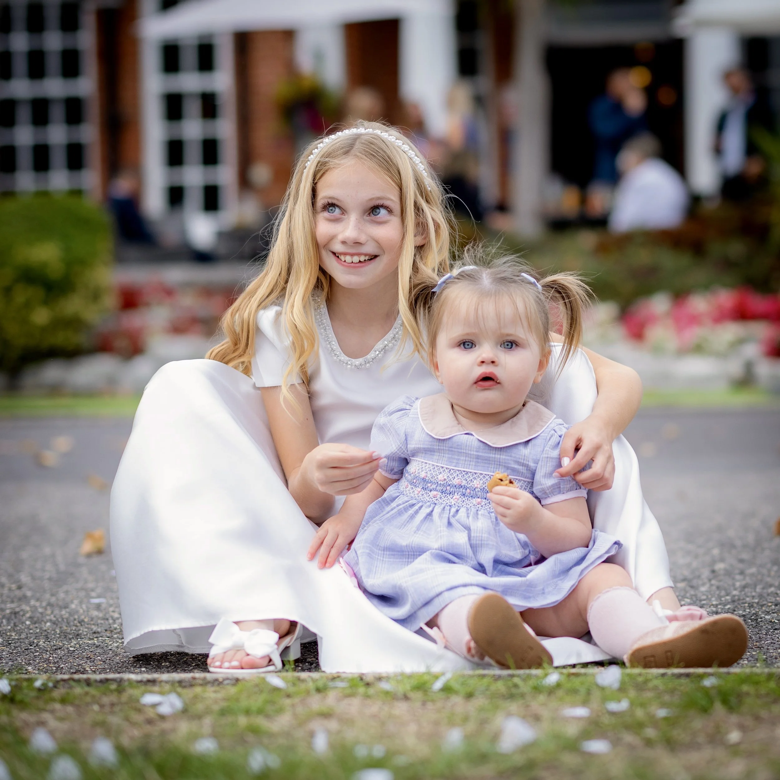 A young girl with blonde hair, blue eyes, and a white pearl headband smiling outdoors, with two younger children, in front of a house with a porch, and a lawn scattered with white flower petals.