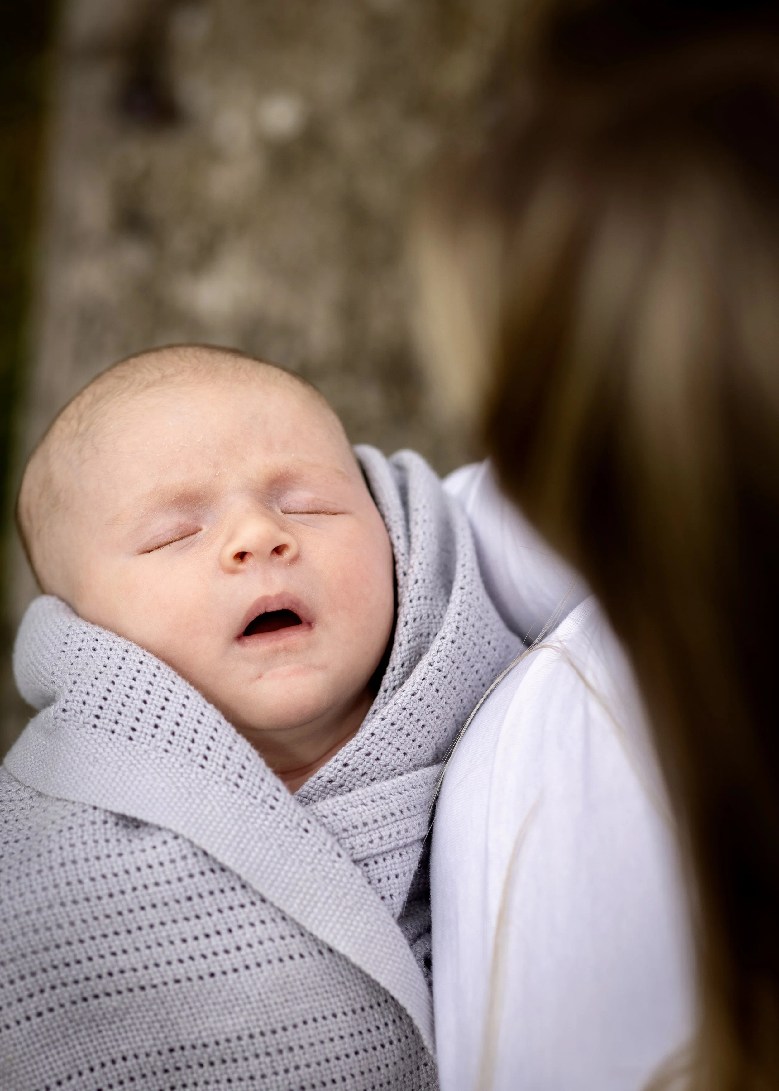 A sleeping baby with light skin and a bald head, dressed in a gray and white knit sweater, being held by an adult.
