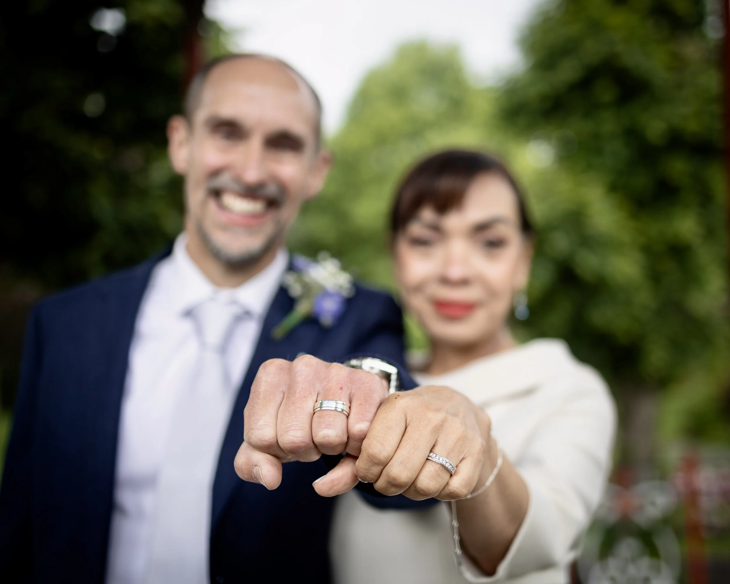 Close-up of a newlywed couple holding hands with wedding rings, both are smiling, outdoors with green trees in the background.