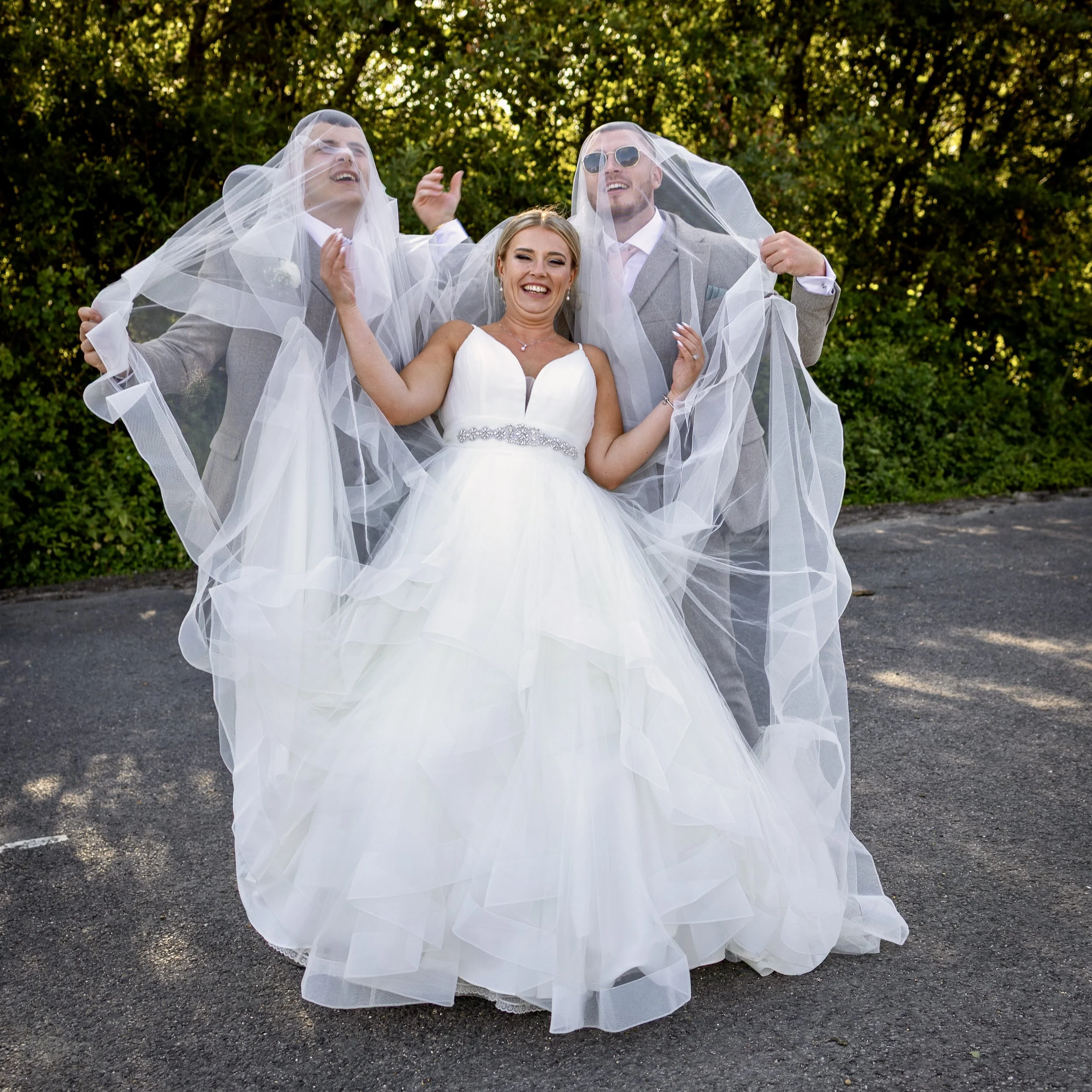 Bride in a white wedding dress smiling, flanked by two grooms in gray suits and sunglasses, outdoors on a paved surface with green trees in the background.