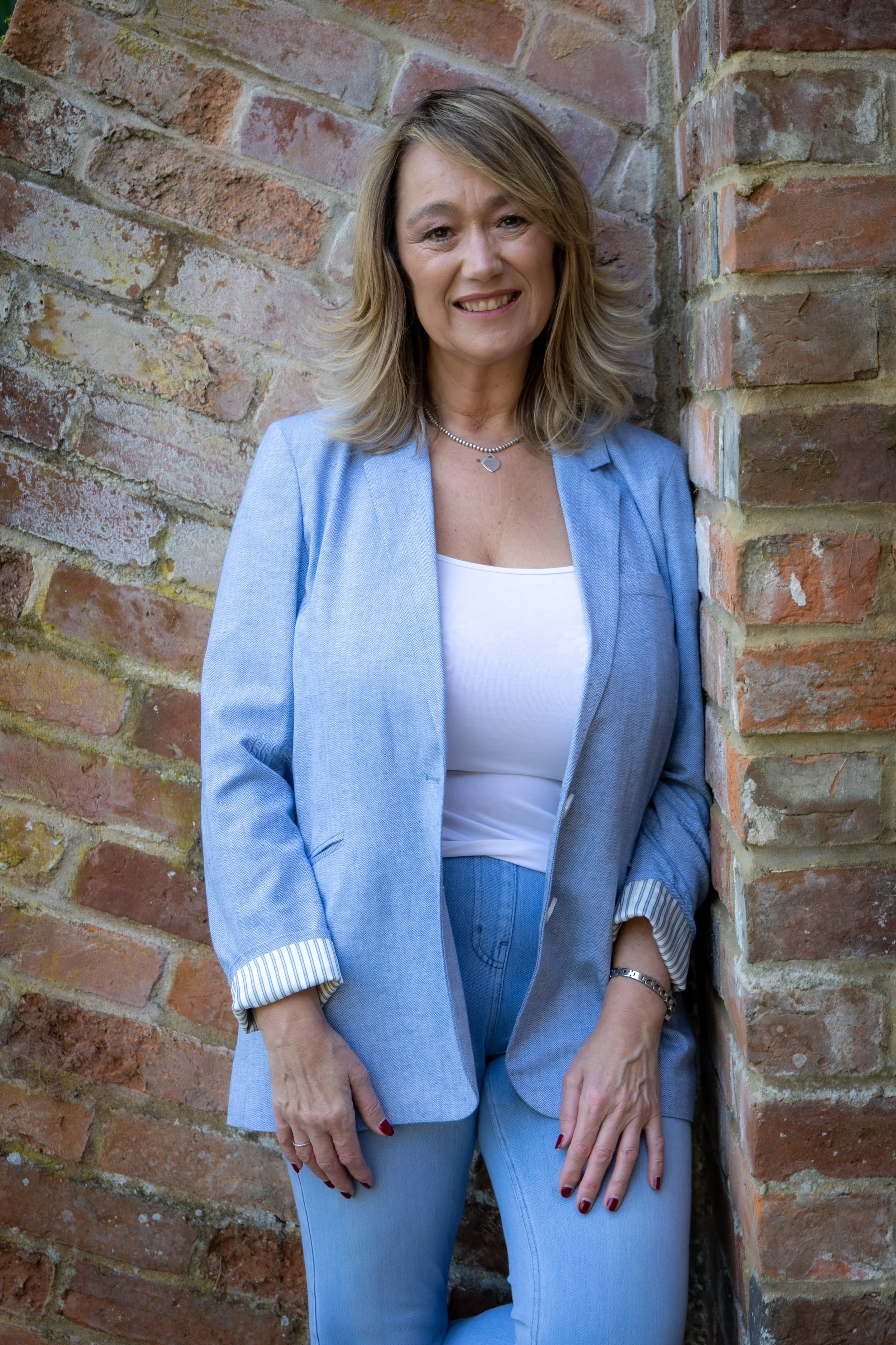 A woman wearing a light blue blazer and jeans standing against a brick wall, smiling at the camera.