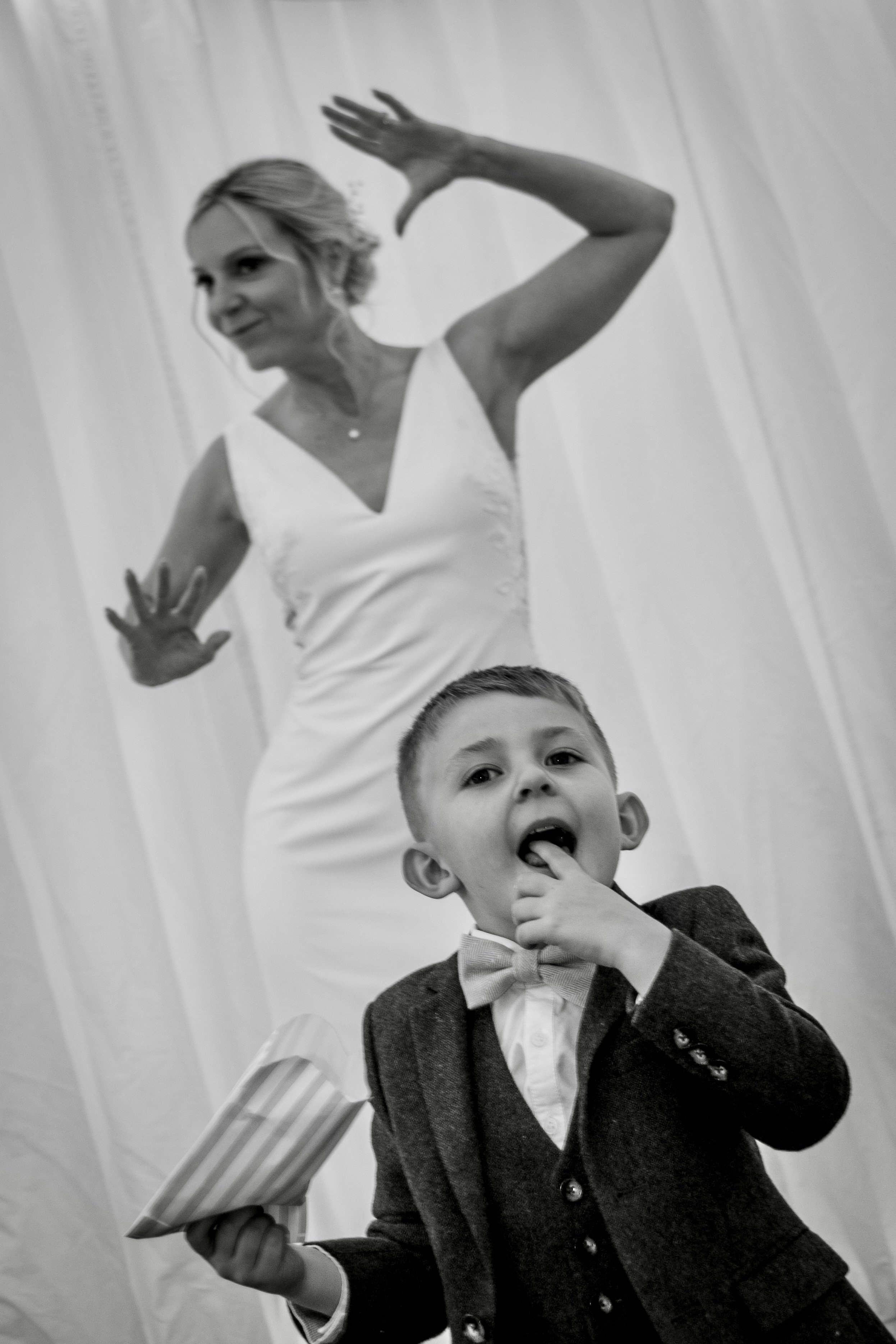 A woman in a white dress poses with her hand raised and fingers splayed, while a young boy in a suit and bow tie stands in front of her, licking his fingers and holding a striped cup.