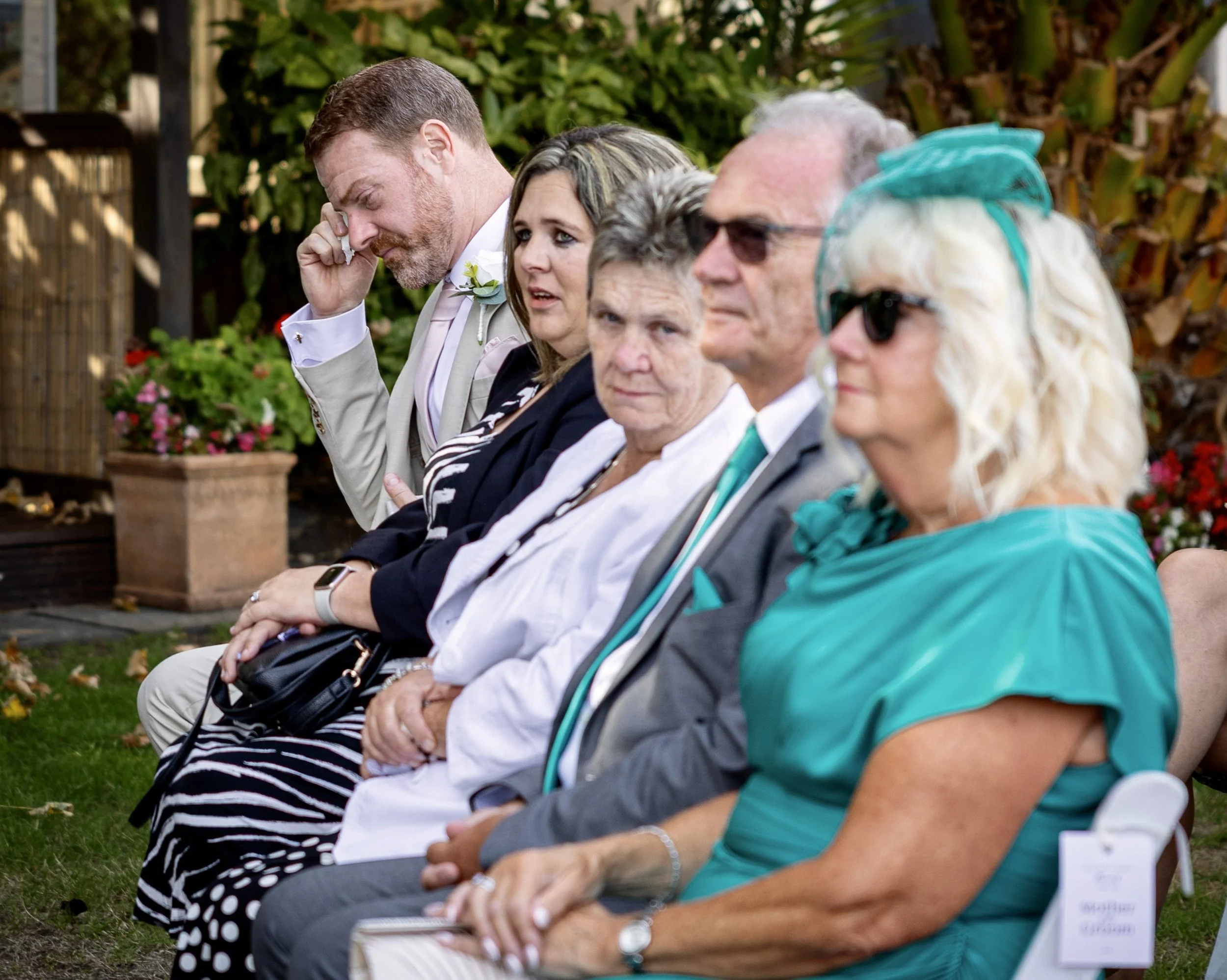 People sitting in a row outdoors during a daytime event, some with sunglasses, with greenery and flowers in the background.