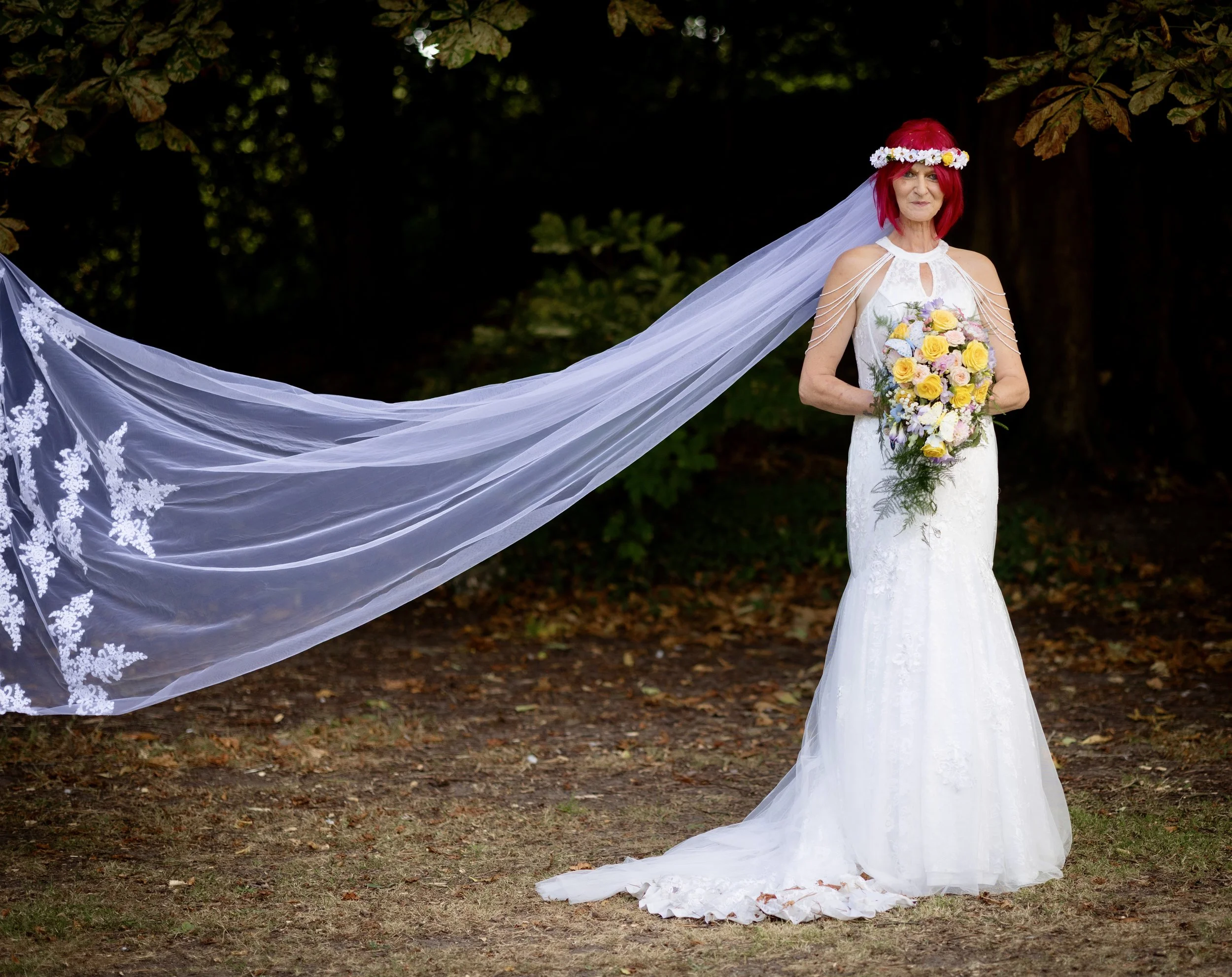 A woman in a white wedding dress with a long veil, holding a bouquet of roses, stands outdoors with a forest background. She has red hair and wears a floral headband.