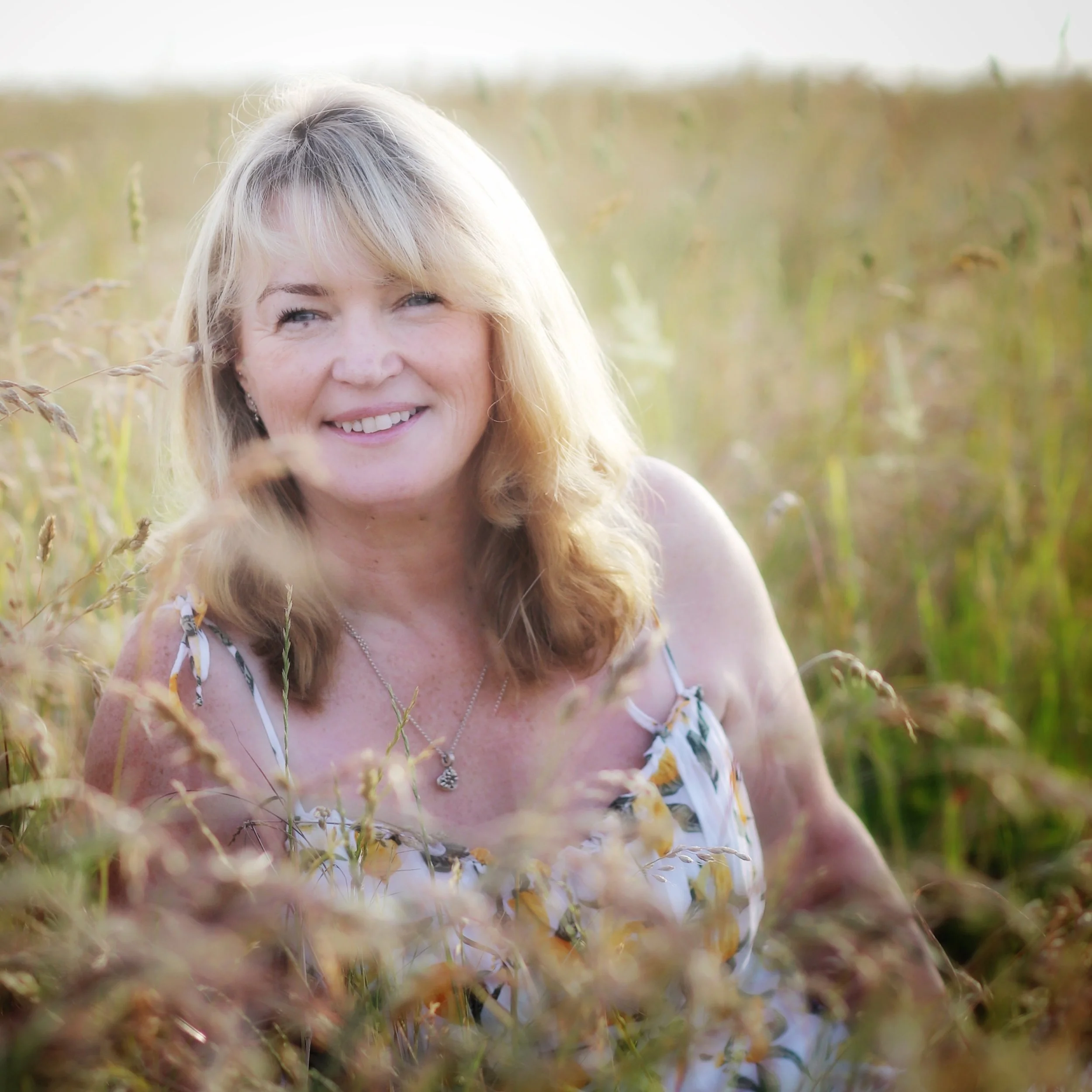 A woman with blonde hair smiling, standing in a field of tall grass or wheat.