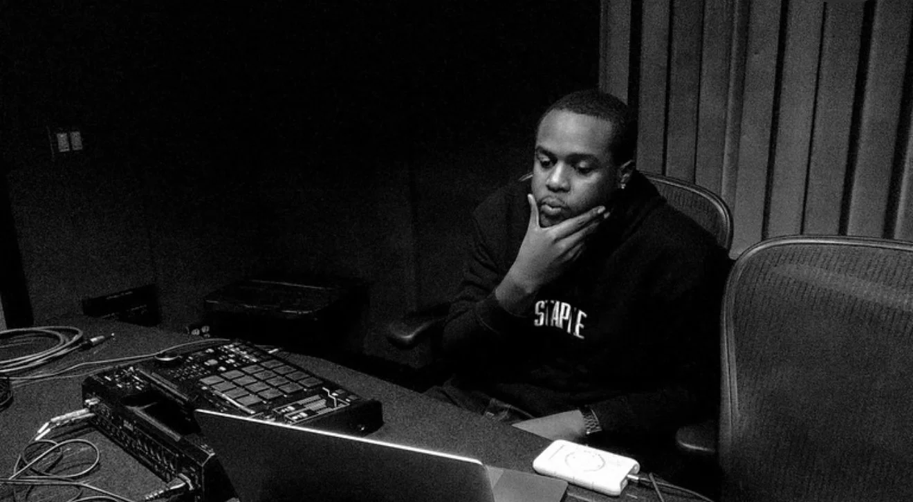 Black Metaphor sitting in a music production studio, looking at a laptop with his hand on his chin, surrounded by music equipment and cables.