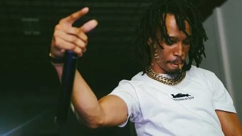 A man with dreadlocks wearing a white T-shirt and layered necklaces, holding a cane or stick, in an indoor setting.