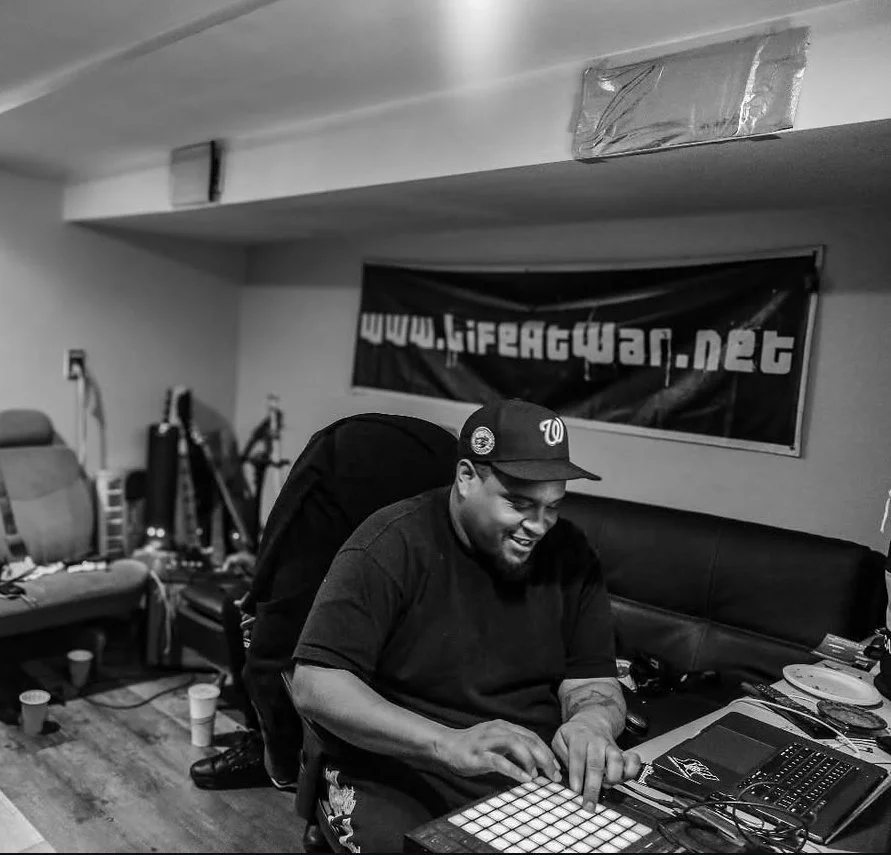 Ceez Neckmusik smiling while working on music production equipment in a room with a banner reading 'www.LifeAttackWar.net' on the wall.