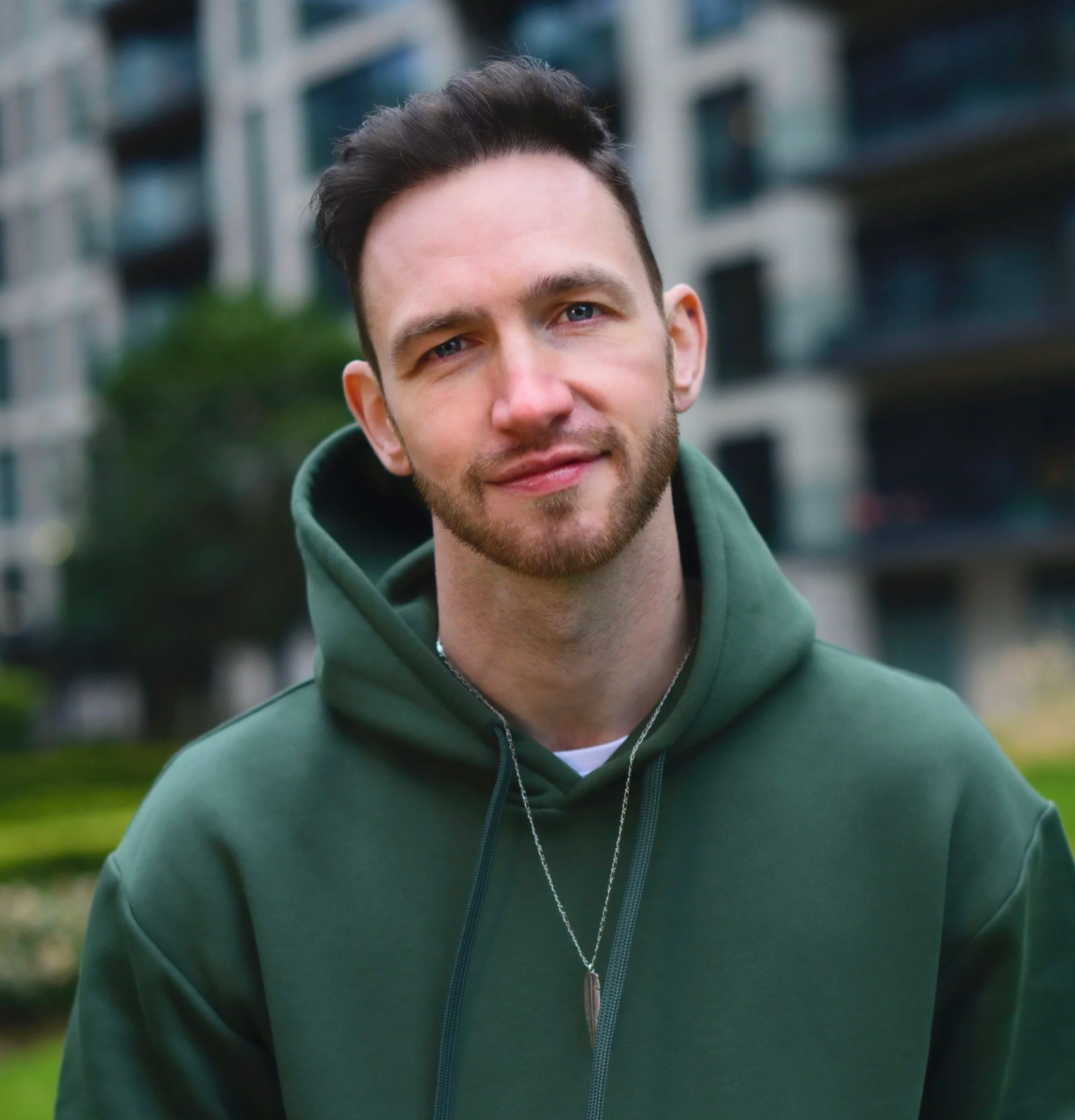 Close-up of a young man with brown hair and blue eyes, smiling with a slight smile, wearing a green hoodie, outdoors with blurry buildings and greenery in the background.