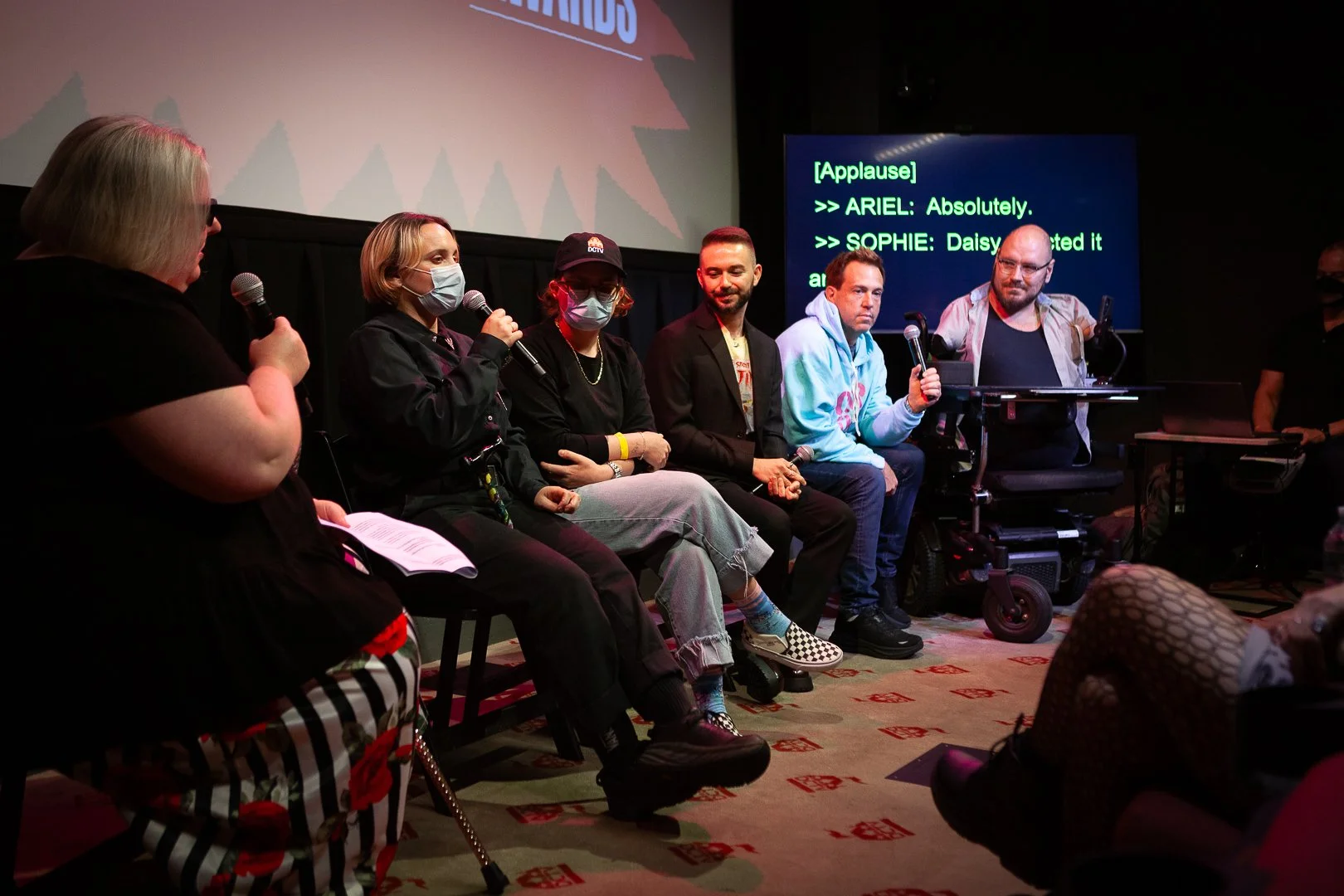 Six event panelists sit in a row speaking to an audience. A screen behind them shows live closed-captioning, and one of the panelists on the right uses a wheelchair.