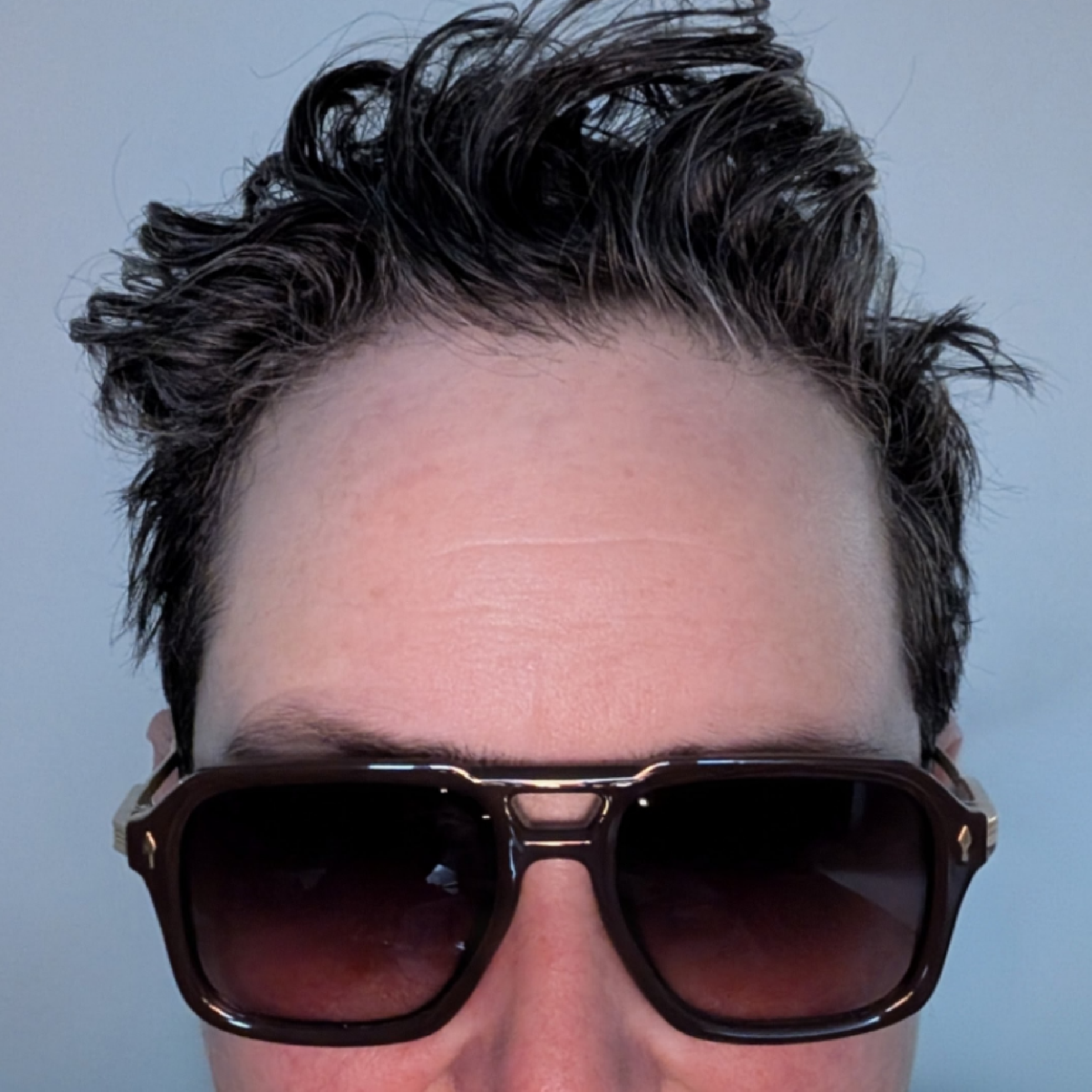 Heather, a person with dark curly hair wears heavy sunglasses. The image cuts off at their nose.