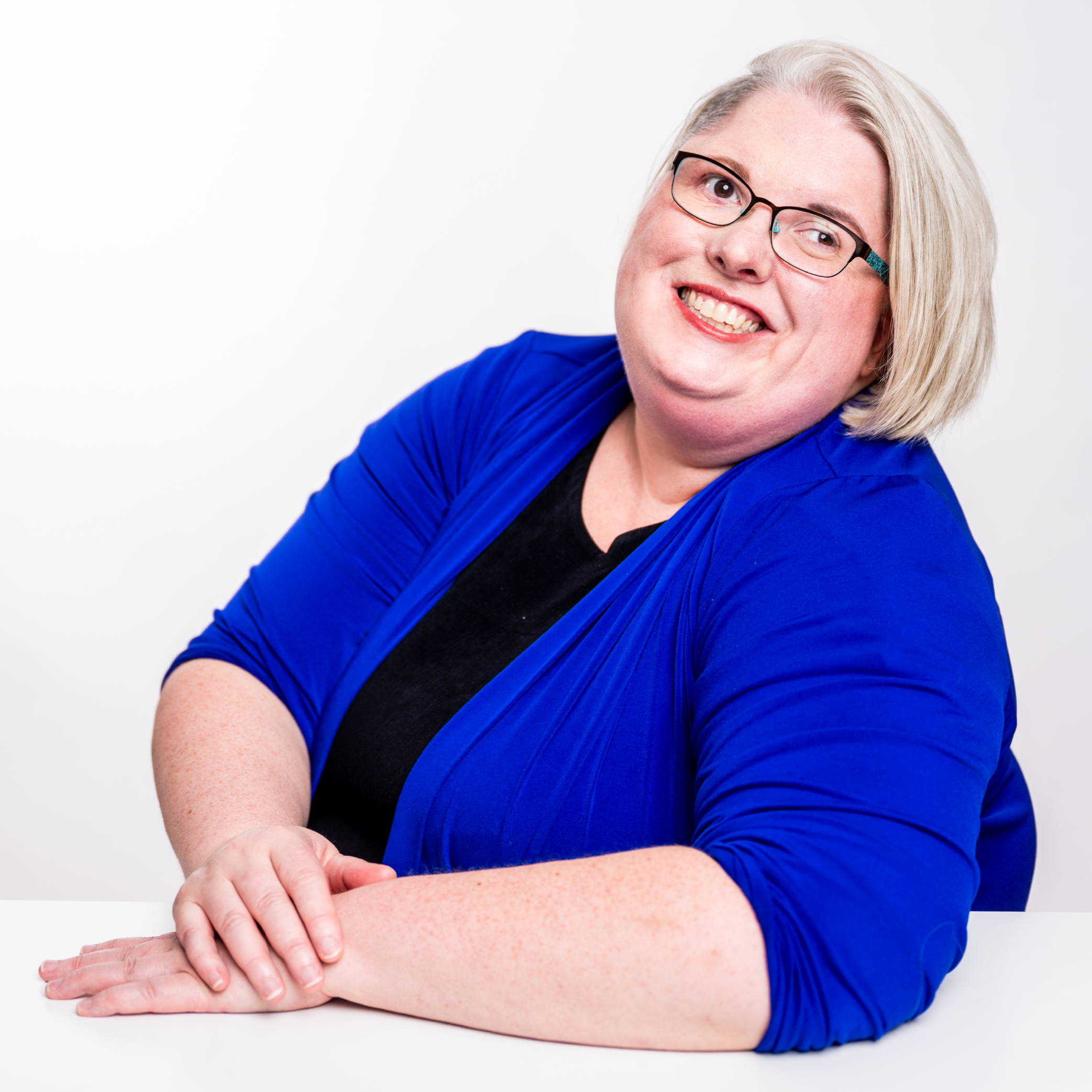 Ariel, a blonde person with glasses, in a brilliant blue top, sits at a white desk smiling and looking up.