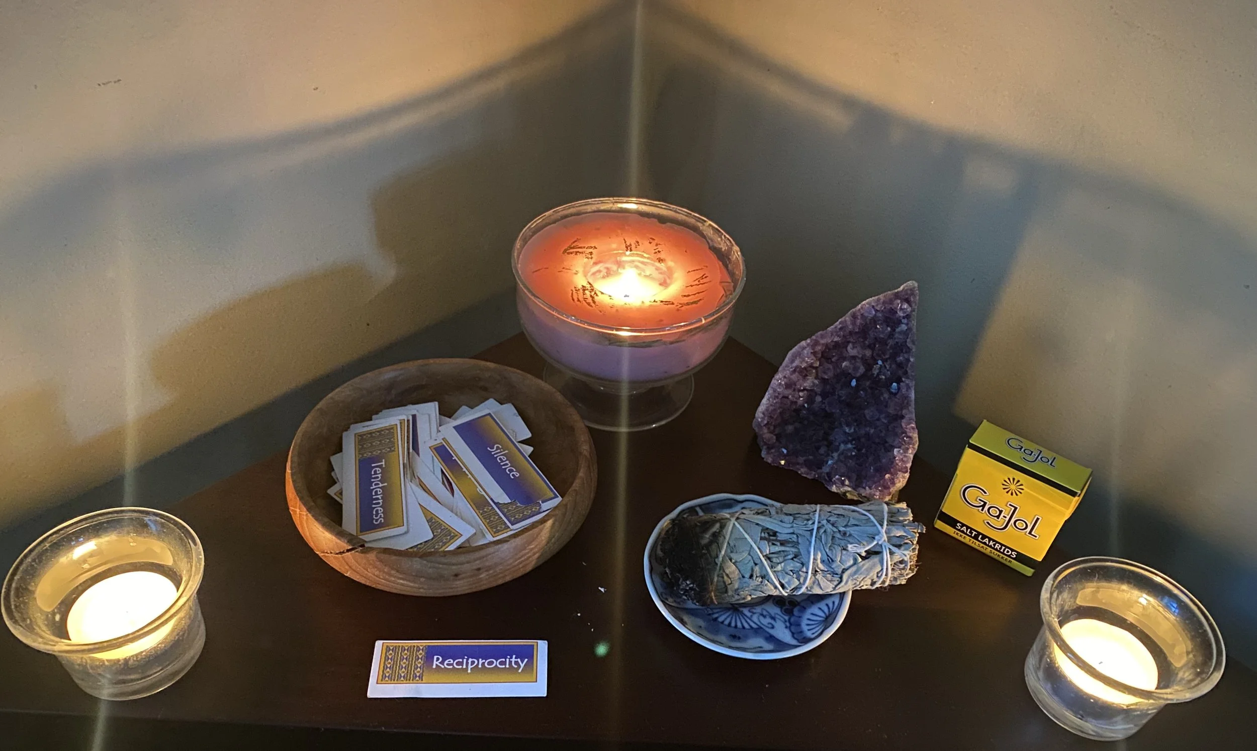 Pink "Abundance" candle between 2 white candles, a wooden bowl of ancestor cards, with one drawn that says "Reciprocity," sage, amethyst, and Icelandic candies