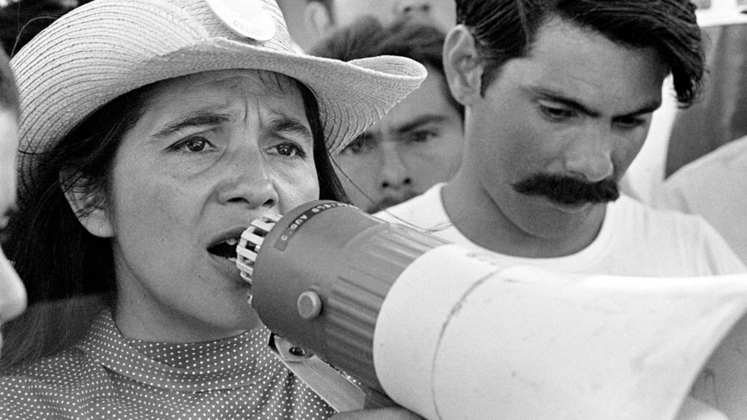 Black and white image of a young Dolores, with a white cowboy hat, speaking into a megaphone beside a man with a mustache. Her face looks clear and determined.