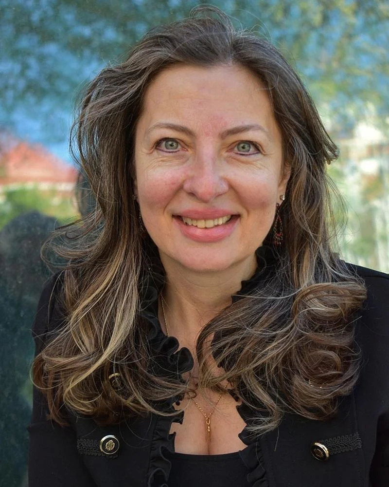 A woman with long wavy brown hair and green eyes, smiling outdoors with a blurred background of trees and sky.
