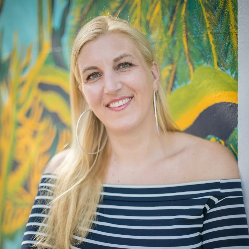 A smiling woman with long blonde hair, wearing large hoop earrings and a navy blue and white striped off-the-shoulder top, standing in front of a colorful abstract mural.