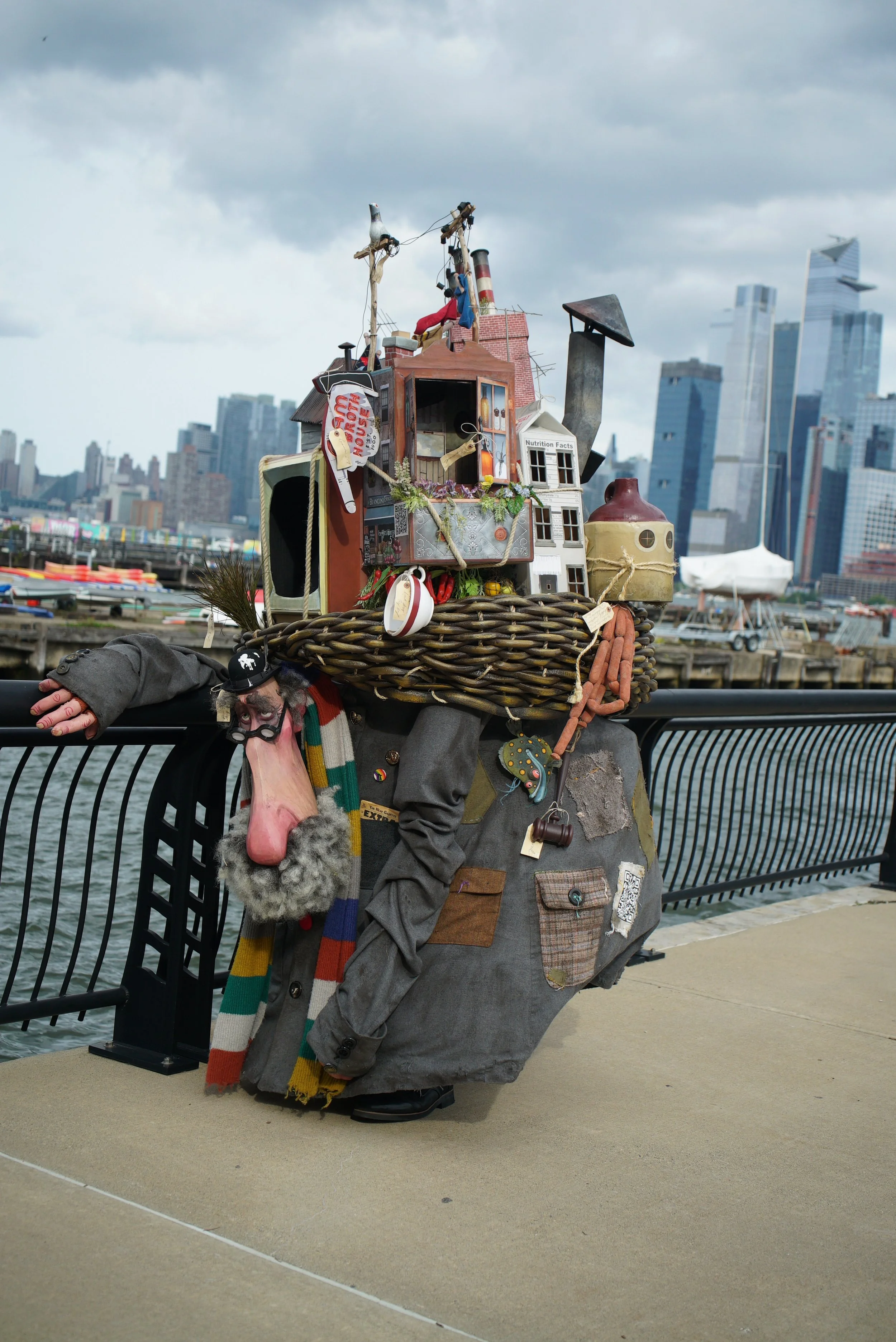 A large puppet with a patchwork suit and colorful scarf, resting on a waterfront promenade with a city skyline in the background. The puppet's head has a long nose, big nose, and bushy eyebrows.