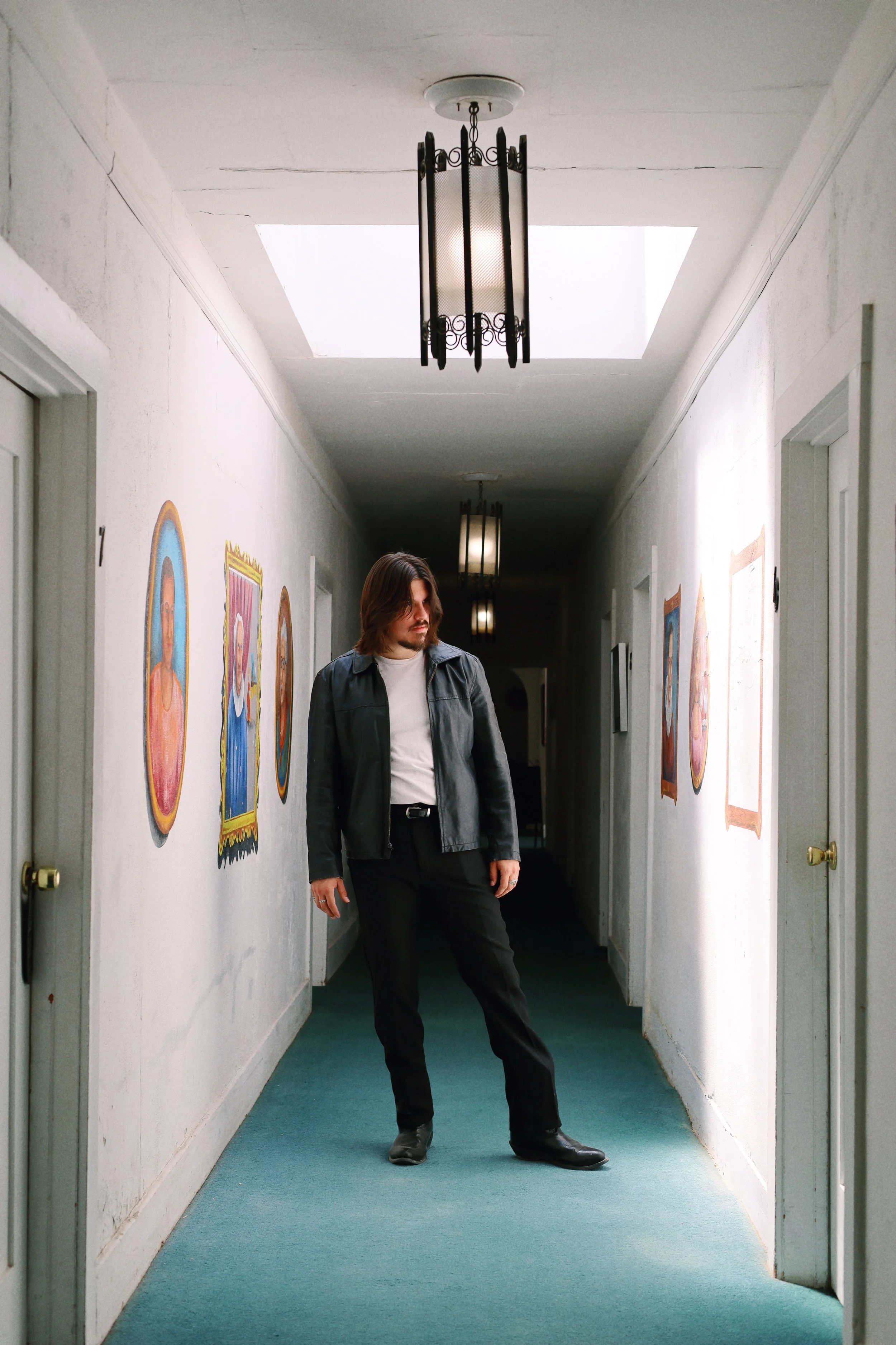 A man with long hair, wearing a black leather jacket and black pants, stands with one foot out in a hallway with white walls and teal carpet, facing a wall with framed pictures.