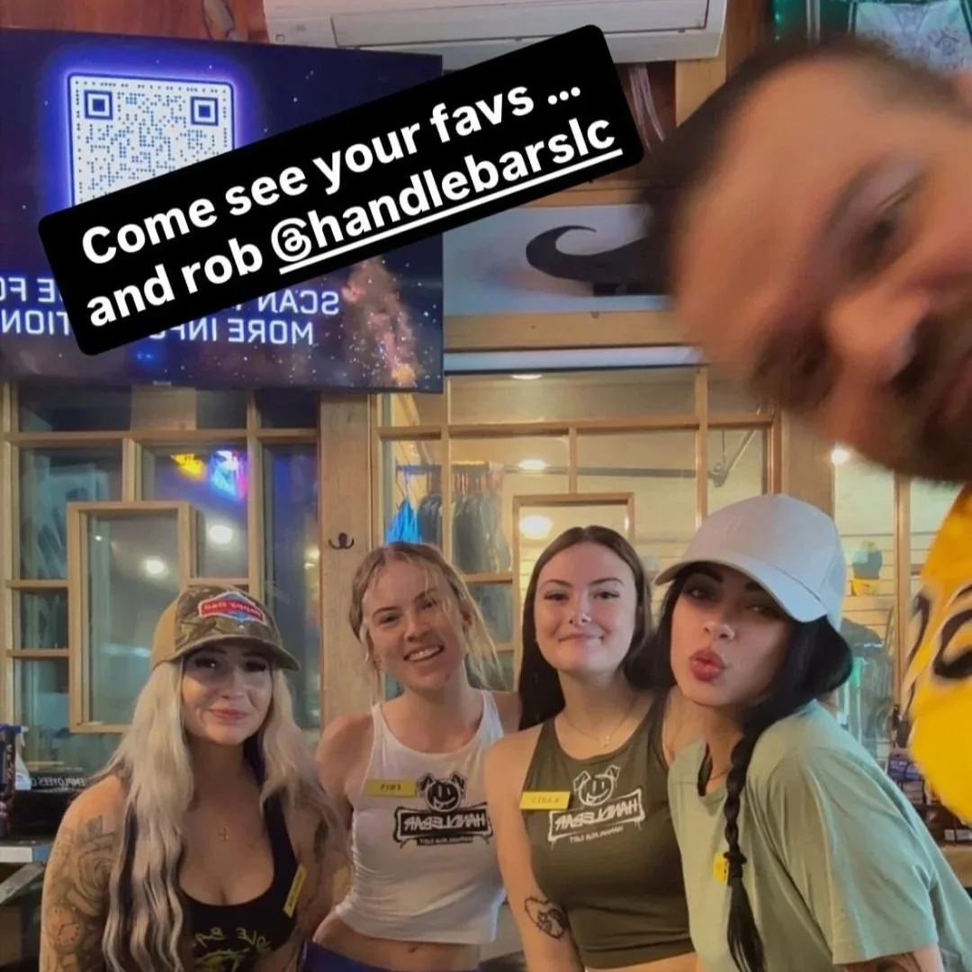 Four women and a man taking a selfie at a bar or cafe, with a digital screen in the background that displays a QR code and the message 'SCAN MORE INFO.' The women are smiling and dressed casually, some wearing tank tops and a baseball cap. The man, p