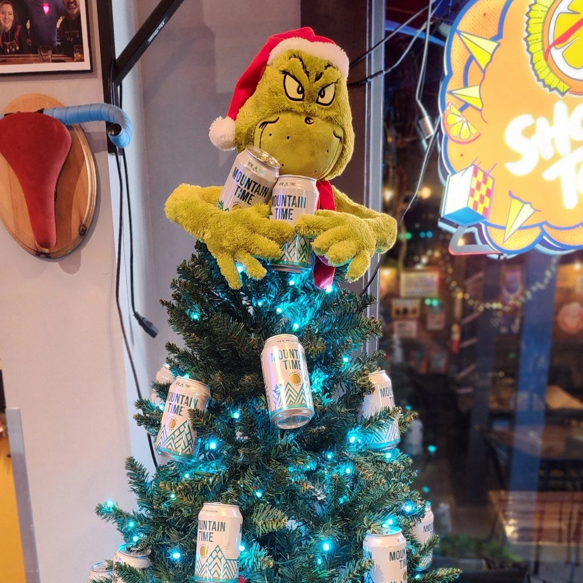 A plush Grinch wearing a Santa hat holding cans of Mountain Time sparkling water, decorated as a small Christmas tree with blue lights, in a store.