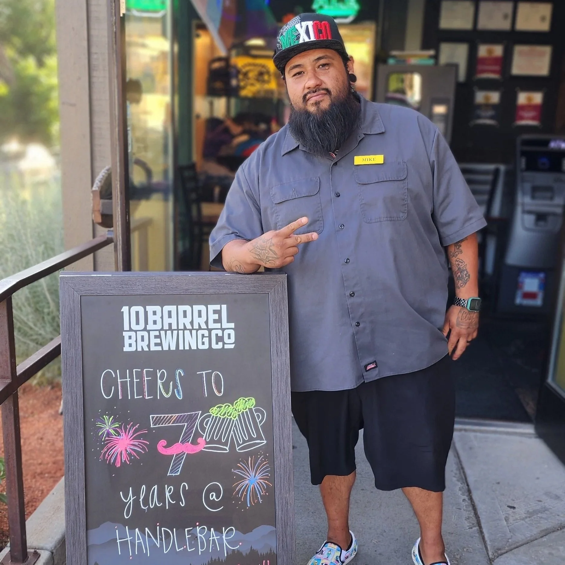 A man with a beard and tattoos wearing a gray shirt, shorts, and a cap points to a chalkboard sign that reads '10 Barrel' and other text, standing in front of a brewing company. The sign has colorful fireworks and beer glass illustrations.