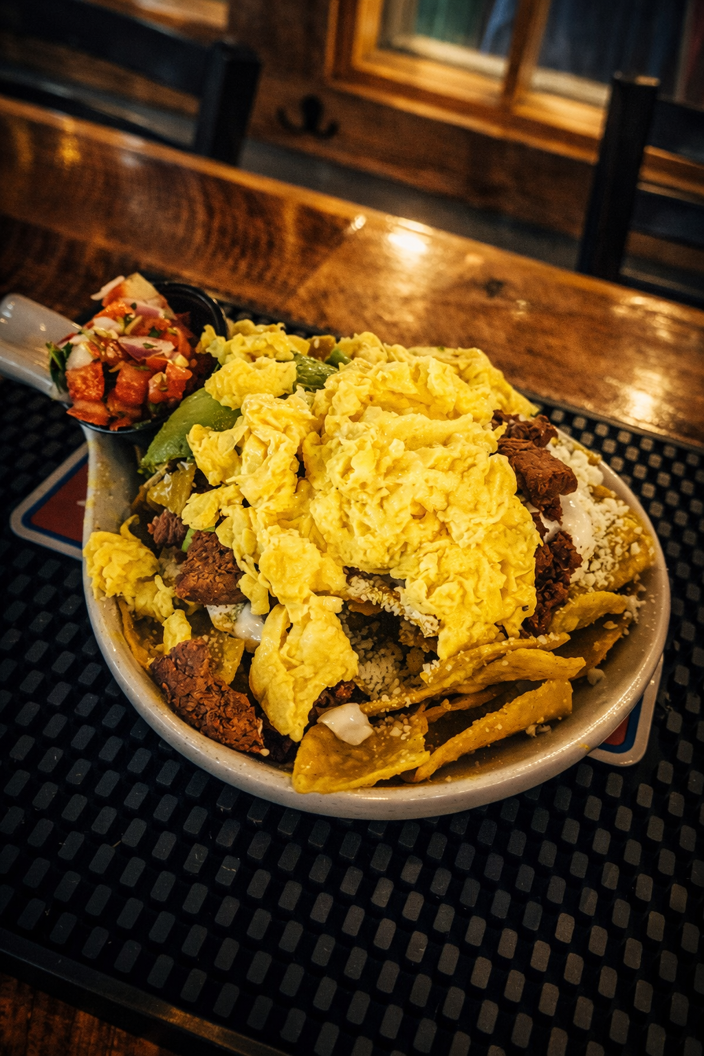 Bowl of tacos with scrambled eggs, seasoned ground beef, shredded cheese, and diced green peppers, garnished with fresh pico de gallo on the side, placed on a black rubber mat on a wooden table.