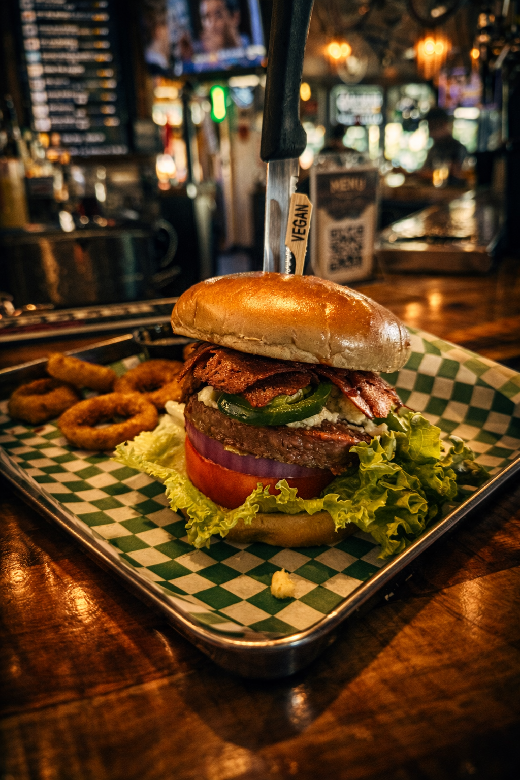 A vegan burger with lettuce, tomato, onion, jalapeños, a vegetable patty, and crispy bacon, served with onion rings on a checkered paper-lined tray in a restaurant.