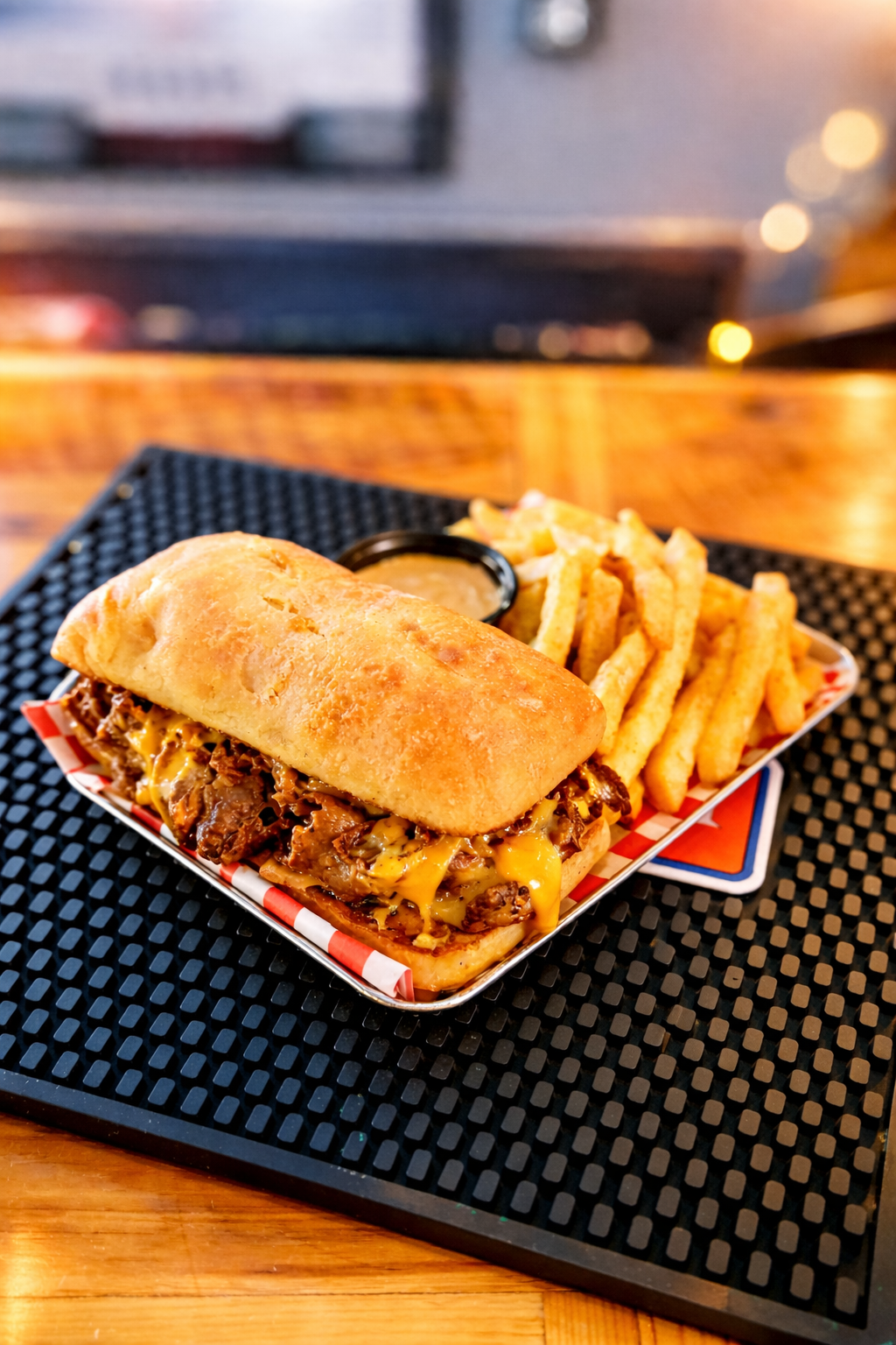 A loaded Philly cheesesteak sandwich with fries and a small cup of dipping sauce on a black textured placemat.
