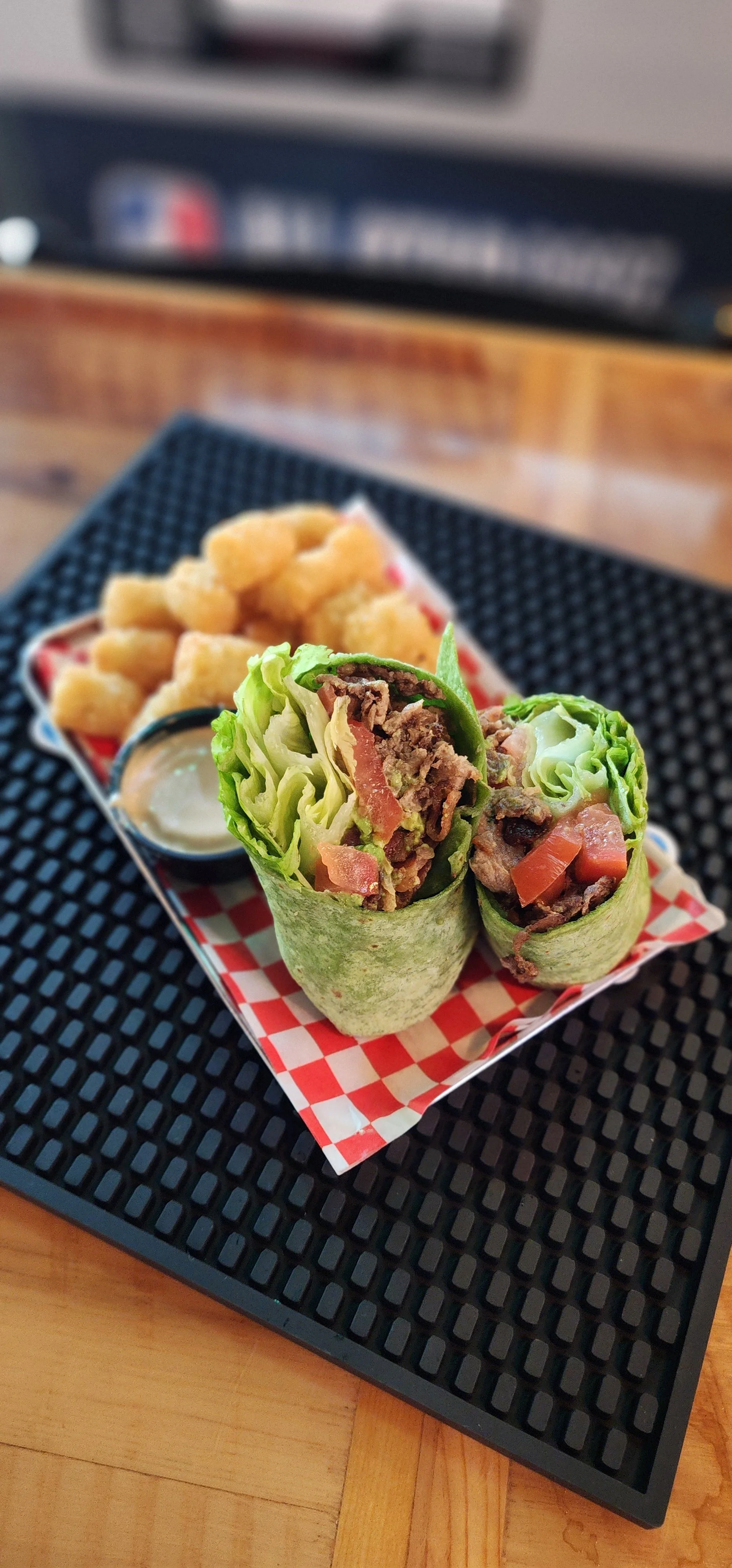 A tray with two beef and vegetable wraps, a small container of dipping sauce, and a side of tater tots on a black textured mat.