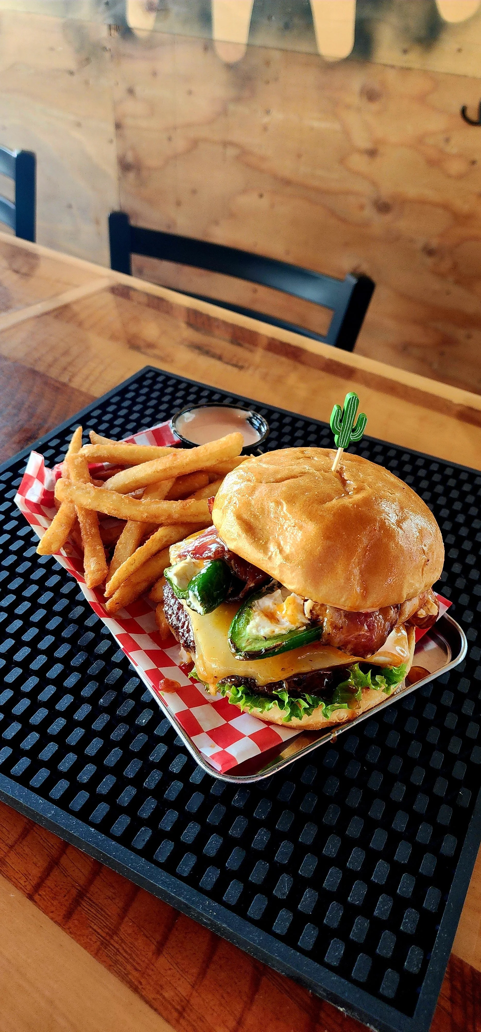 A burger with jalapeños, lettuce, cheese, and bacon, served with a side of French fries and dipping sauce on a black tray.